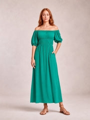 Model in a teal off-shoulder midi dress with voluminous half-length balloon sleeves, gathered waist, and flowing A-line silhouette, posed with one hand in a pocket.