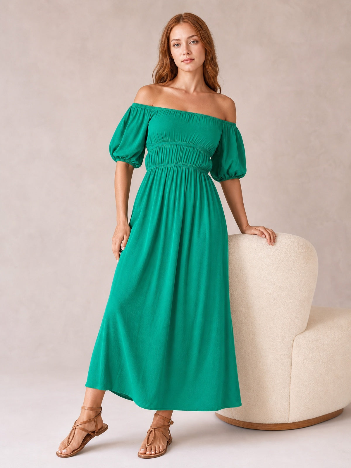 Model in a vivid teal off-shoulder midi dress with gathered waist, voluminous half-length balloon sleeves, and a flowing A-line silhouette, posing beside a chair.