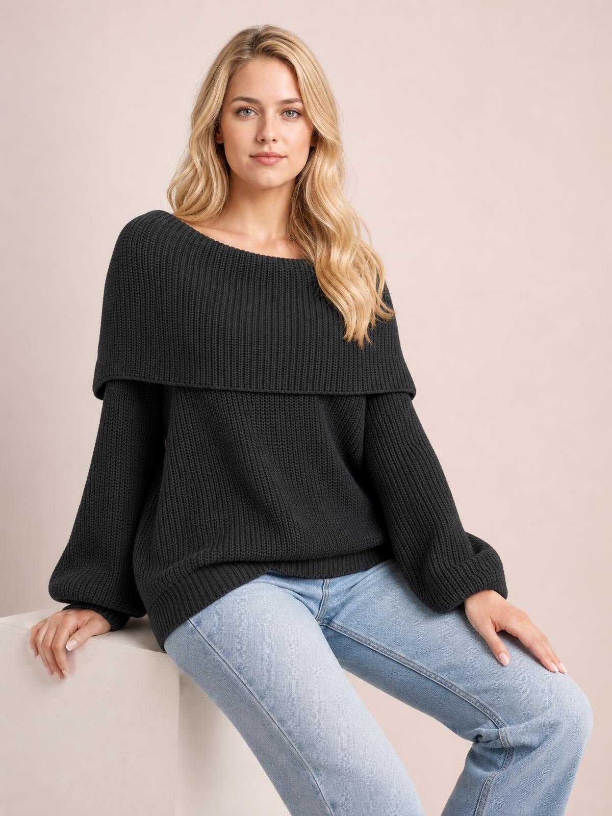 Seated model wears a black off-shoulder long sleeve sweater with an oversized silhouette, wide neckline, ribbed knit construction, and relaxed fit.