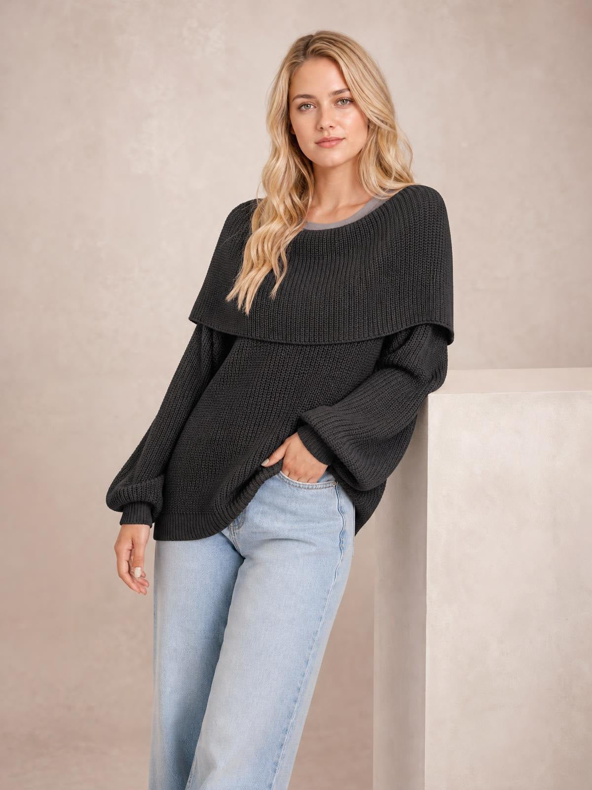 Model wears a black oversized ribbed long sleeve sweater with a wide neckline, cape-like yoke, relaxed silhouette, and ribbed cuffs while leaning with one hand in her jeans pocket.