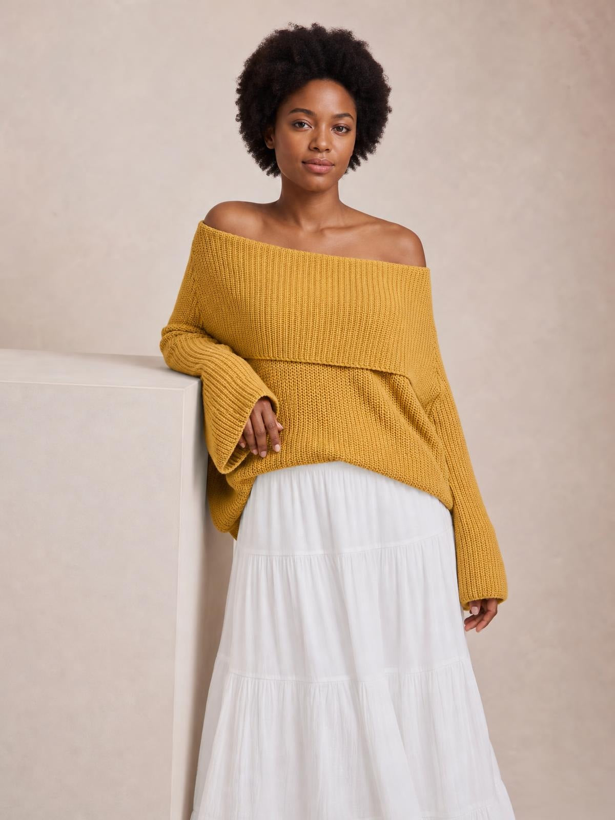 Model leans against a ledge wearing a mustard-yellow oversized ribbed off-shoulder long sleeve sweater with a relaxed silhouette and extended cuffs.