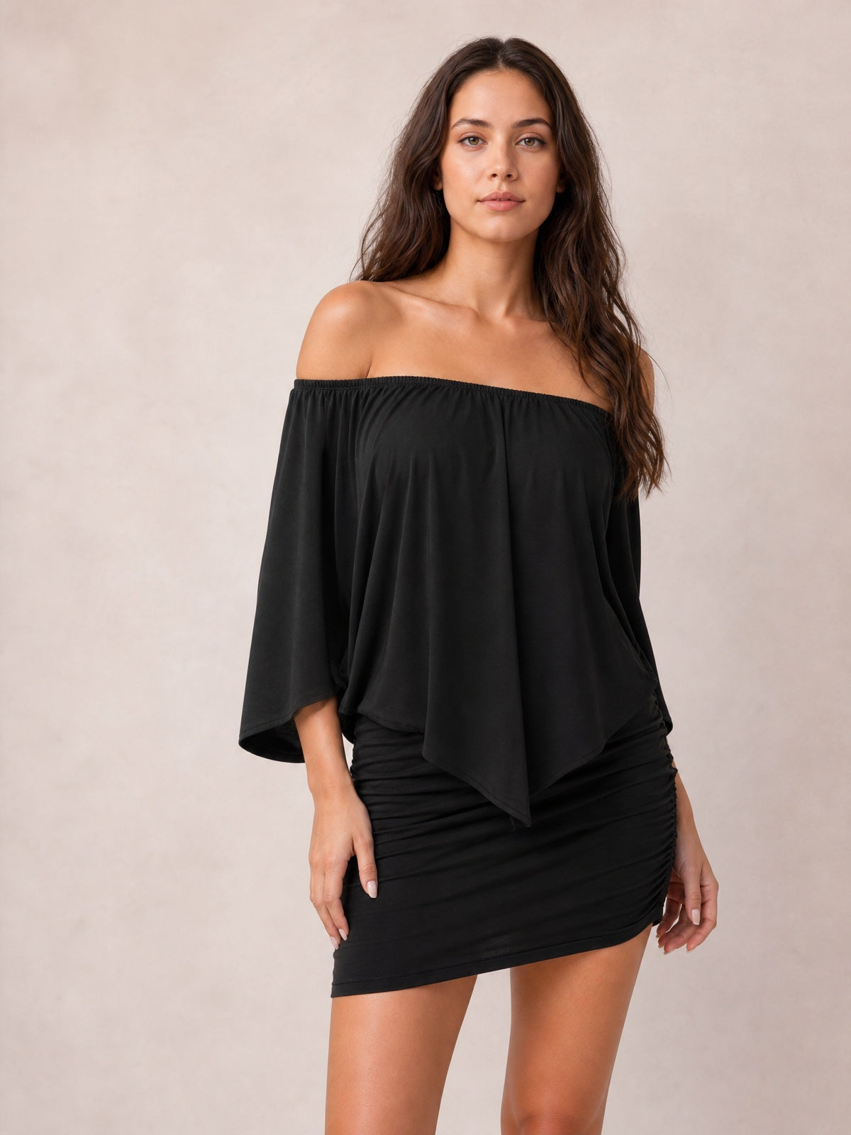 Model poses in a black off-shoulder mini dress with three-quarter sleeves, a softly draped top, and a gathered, ruched skirt.