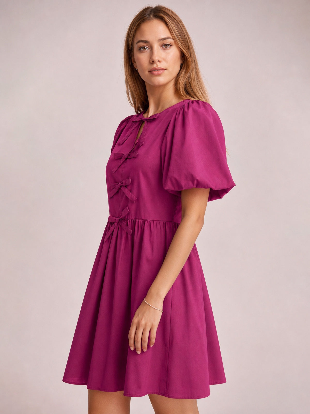 A model stands in a cerise bow ruched mini dress with voluminous puff sleeves, gathered waist, front bow details, and a gently flared silhouette.