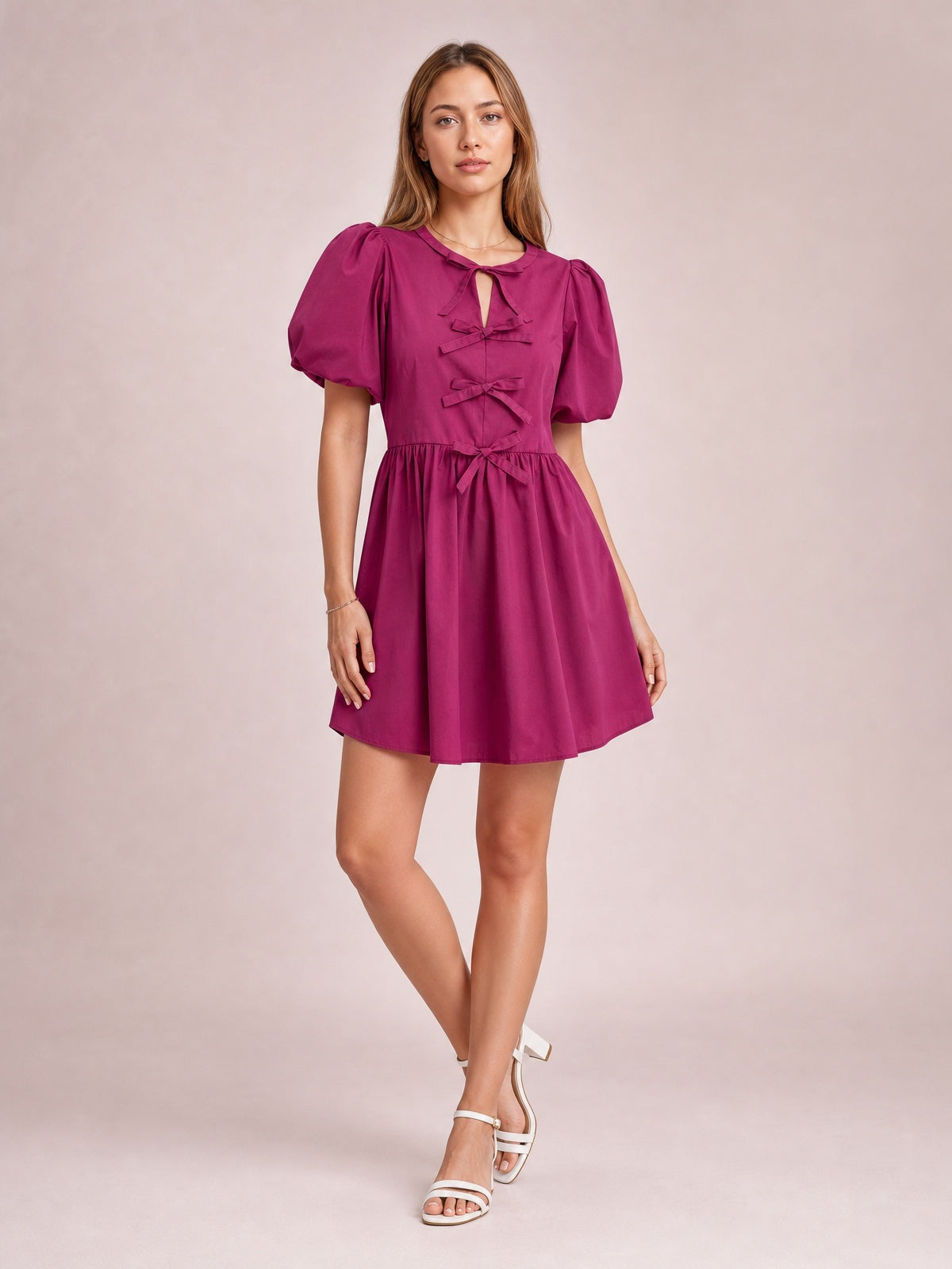 Model poses in a cerise bow ruched mini dress with voluminous puff sleeves, a gathered waist, multiple front bows, and a gently flared skirt.