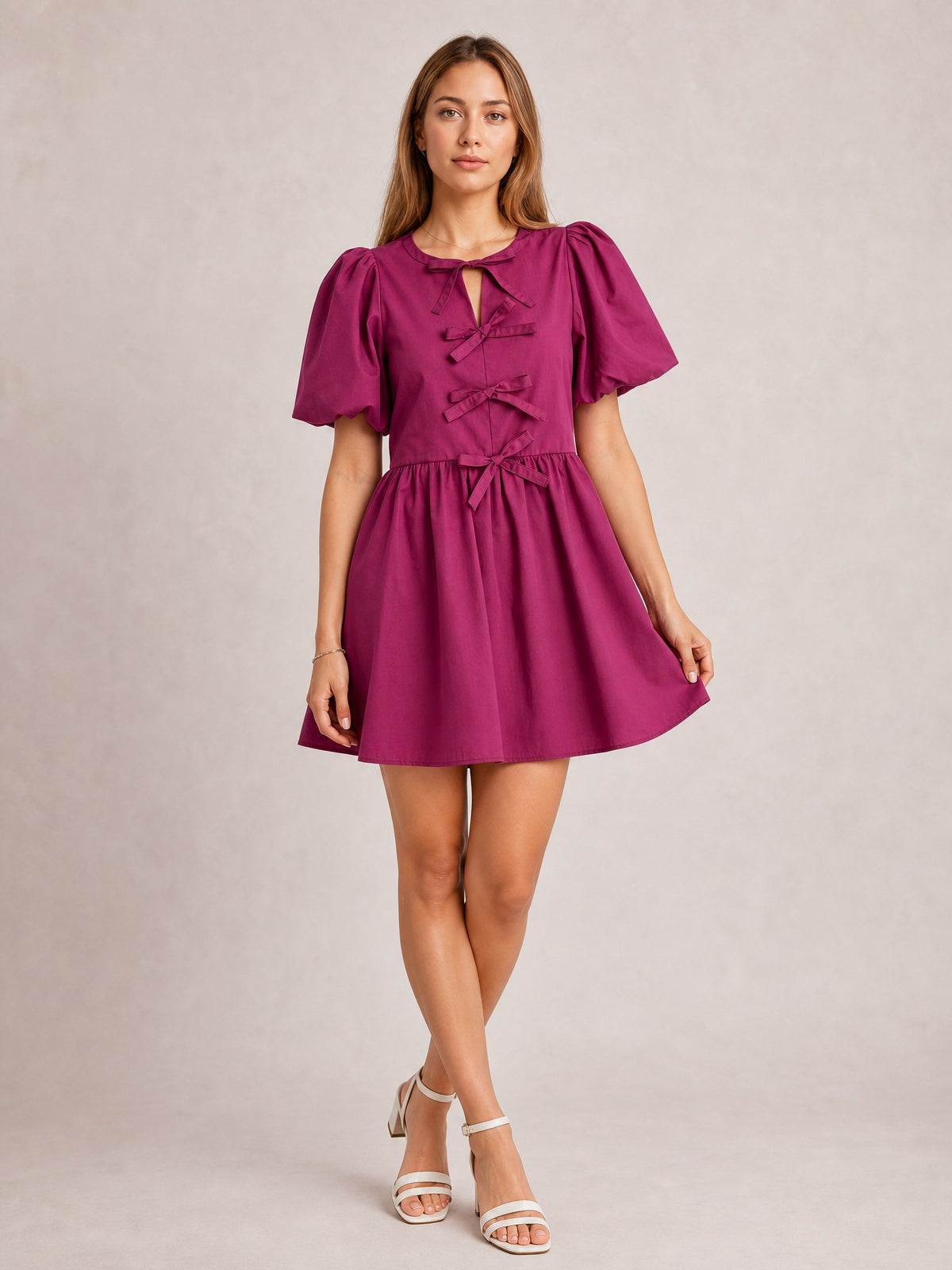 Model poses in a cerise bow ruched mini dress with voluminous puff sleeves, a softly gathered waist, multiple front bows, and a gently flared skirt.