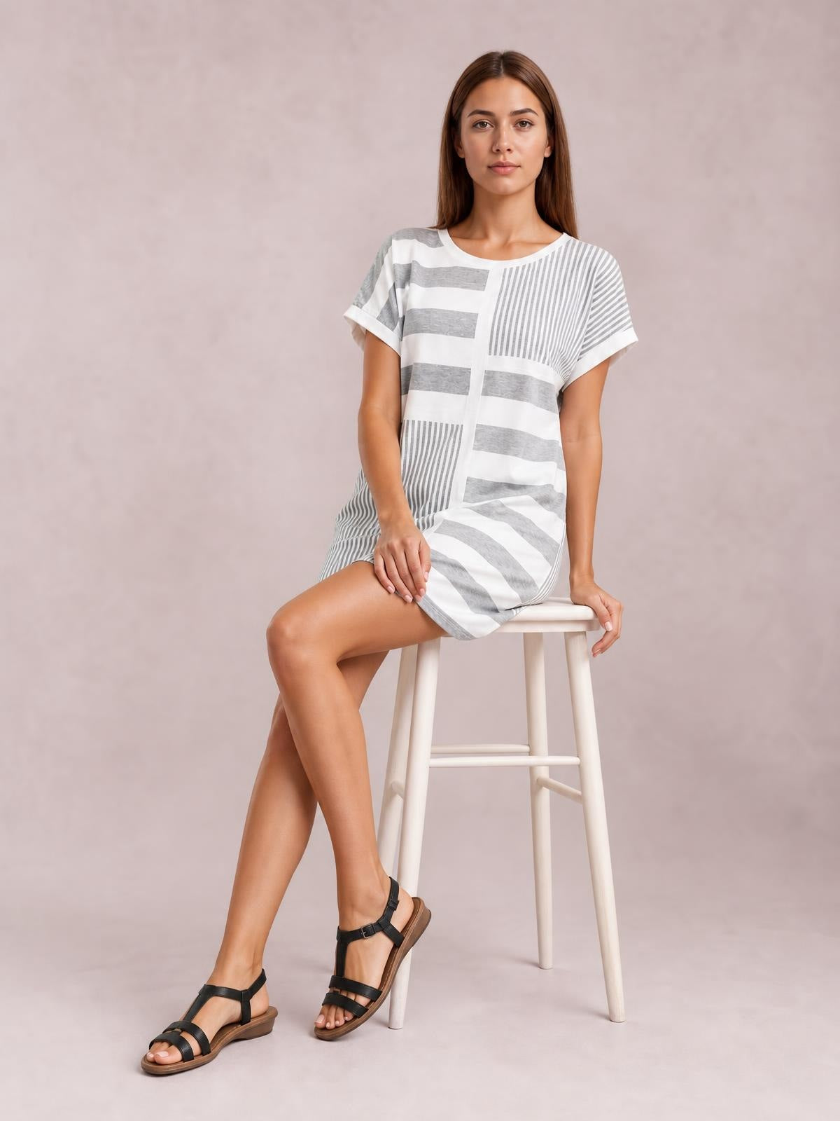 Model seated in a relaxed pose wearing a white and gray striped round-neck mini tee dress with short sleeves and a straight silhouette.