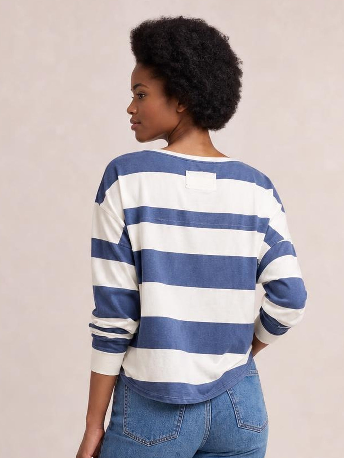 Model shown from behind wearing a relaxed blue-and-white contrast striped long-sleeve T-shirt with a round neck, exposed seams, and curved hem.