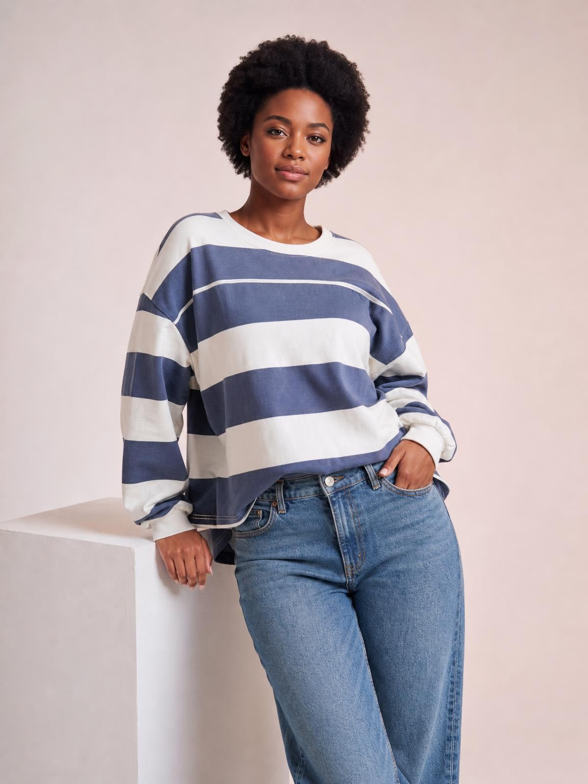 A model leans against a pedestal wearing a relaxed blue and white contrast striped long-sleeve T-shirt with a round neck and exposed seam details.