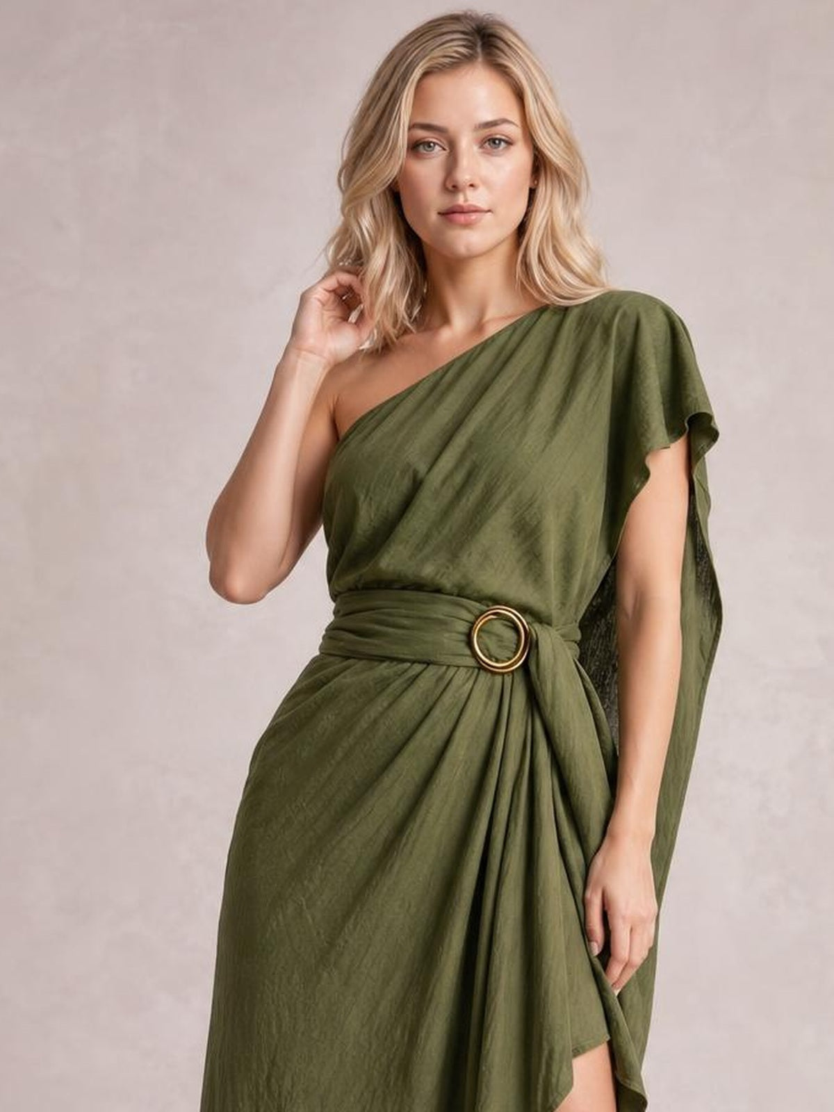 A model poses in an olive-green one-shoulder draped maxi dress with gathered waist, circular buckle, flowing silhouette, and asymmetrical cape-like sleeve.
