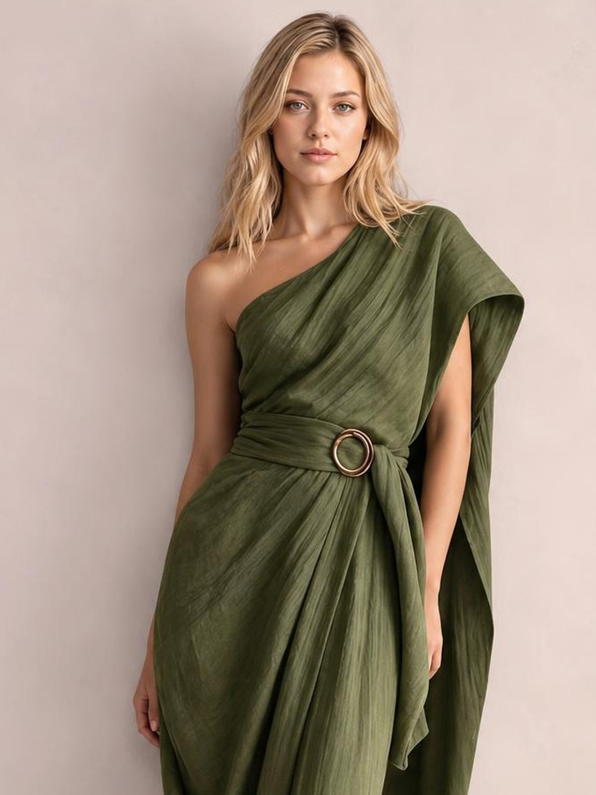 Model poses in a one-shoulder olive green draped maxi dress with a gathered waist, circular buckle, flowing pleats, and asymmetric cape-like sleeve.