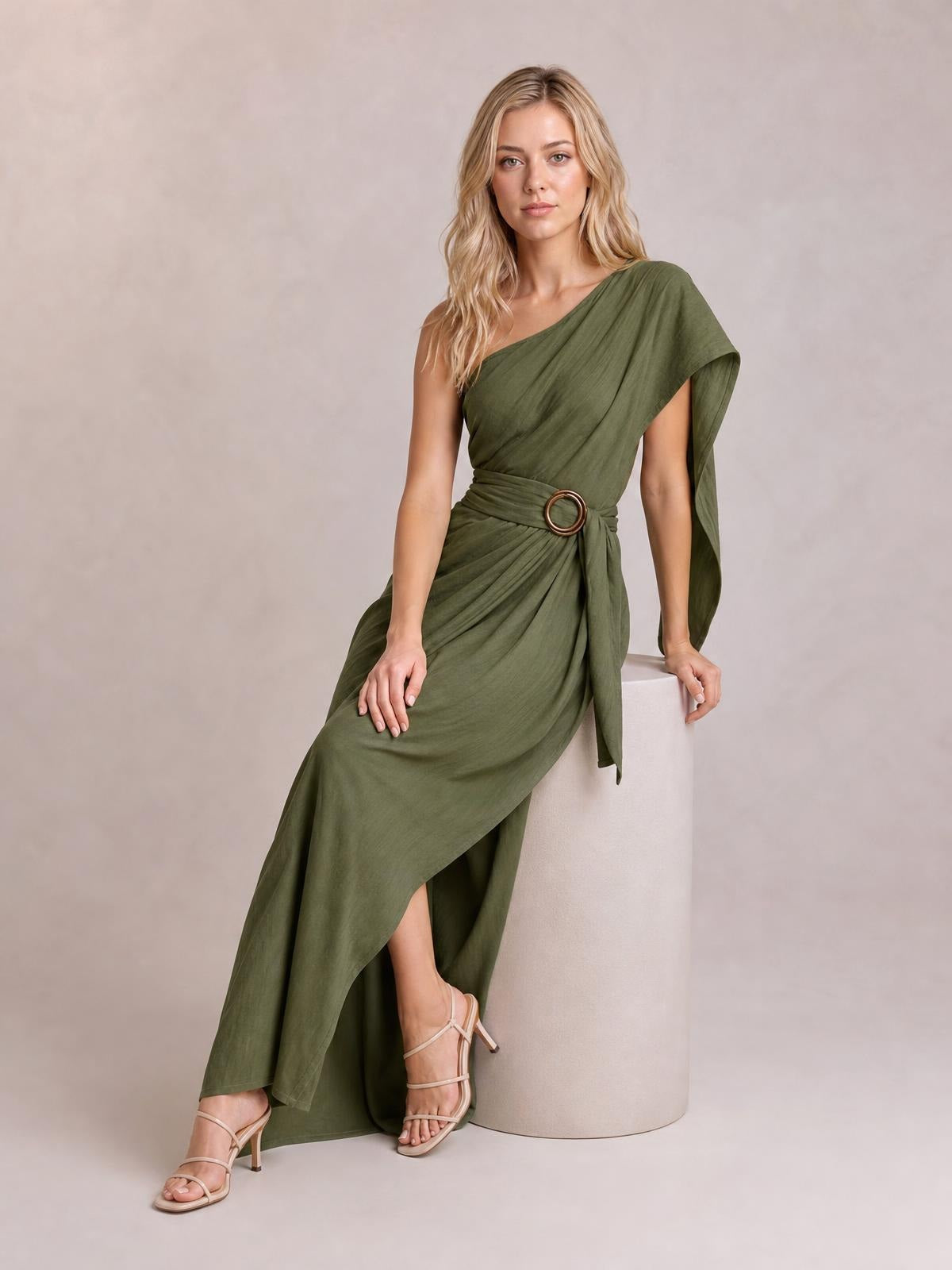Model seated in an olive green one-shoulder draped maxi dress with a waist buckle, gathered fabric, asymmetrical hem, and flowing side sash.