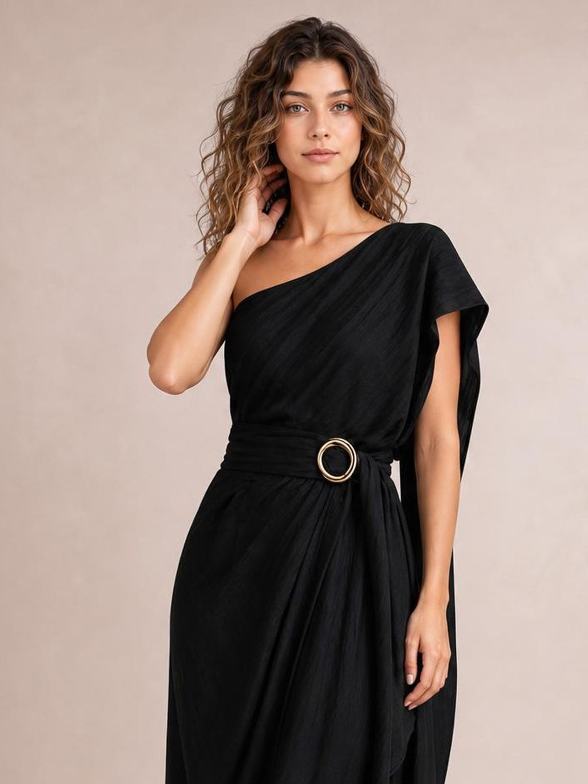 A model poses in a black one-shoulder draped maxi dress with an asymmetrical neckline, gathered waist, circular buckle, and flowing silhouette.