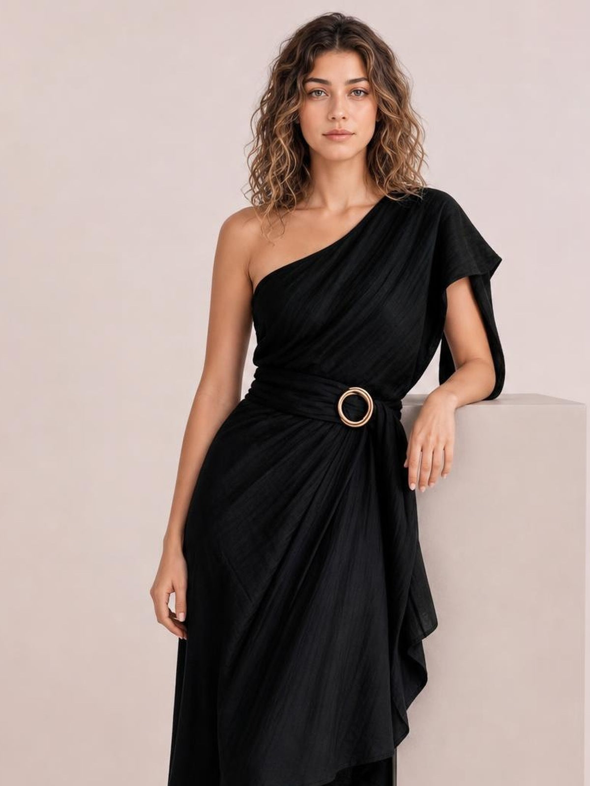 A model leans against a surface wearing a black one-shoulder draped maxi dress with a gathered waist, circular buckle, and flowing asymmetric skirt.