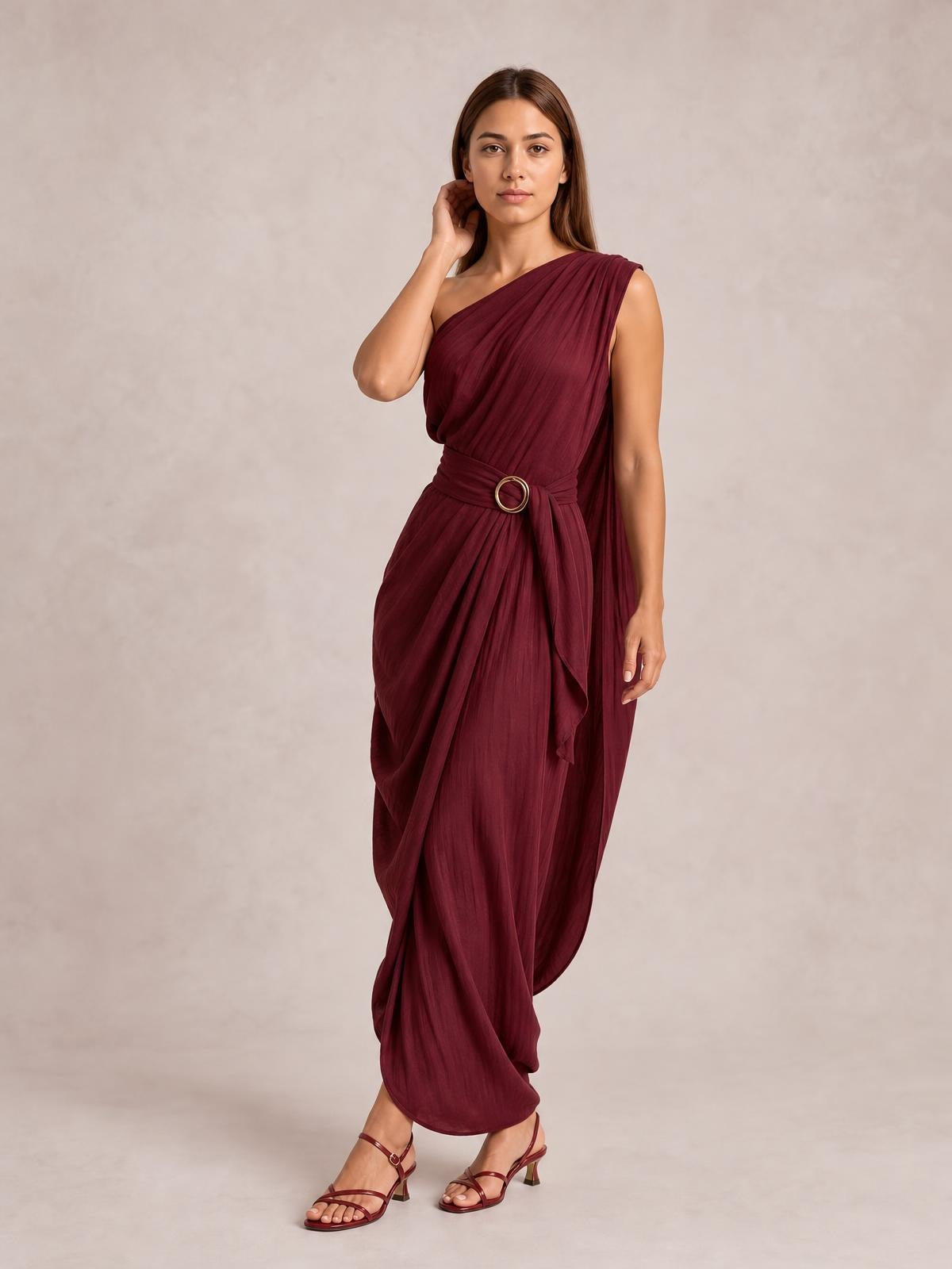 Model poses in a burgundy one-shoulder draped maxi dress with gathered fabric, a defined waist, circular buckle, and flowing asymmetric silhouette.