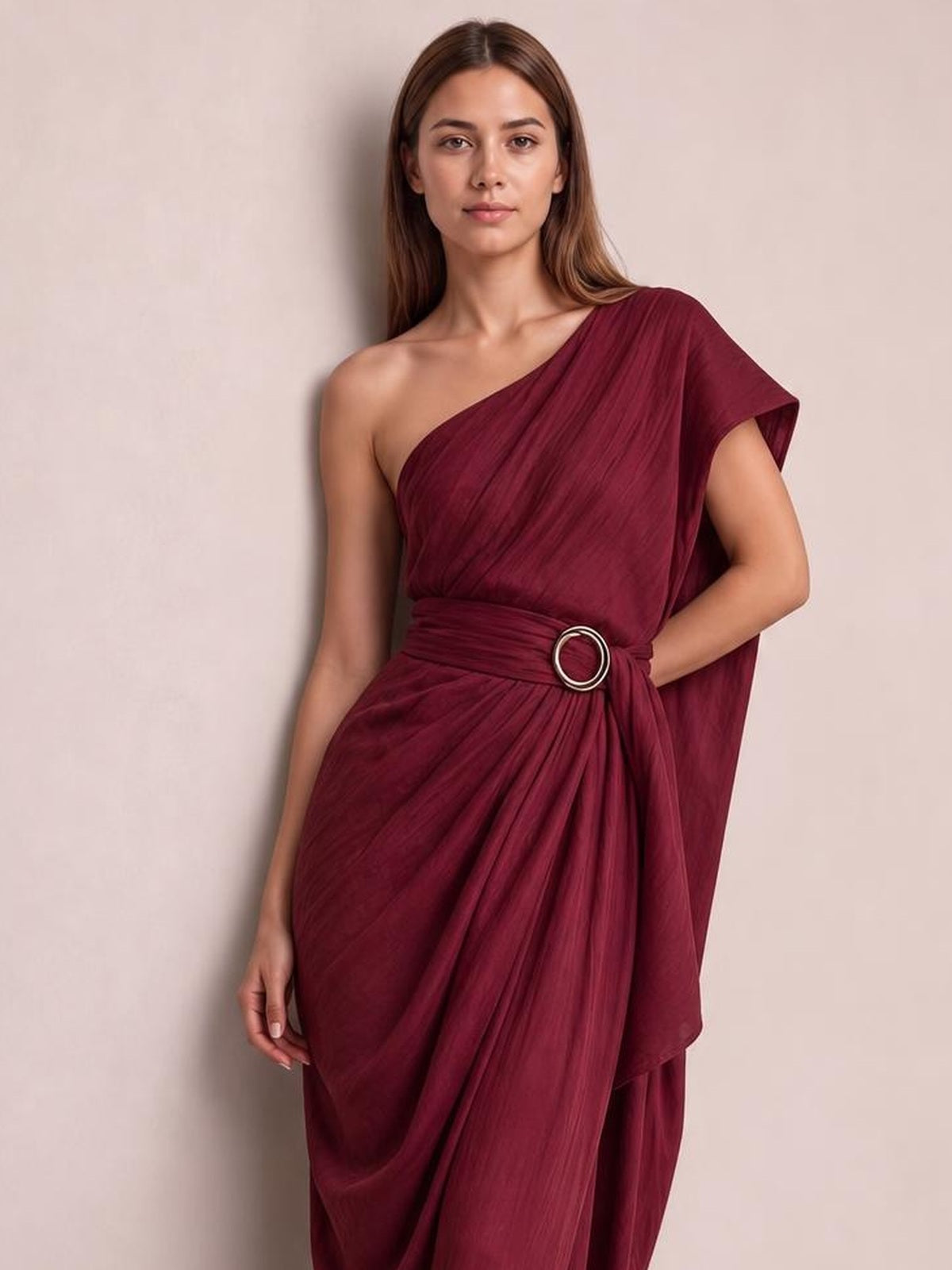 Model poses in a burgundy one-shoulder draped maxi dress with a gathered waist, circular buckle, and flowing pleated silhouette.