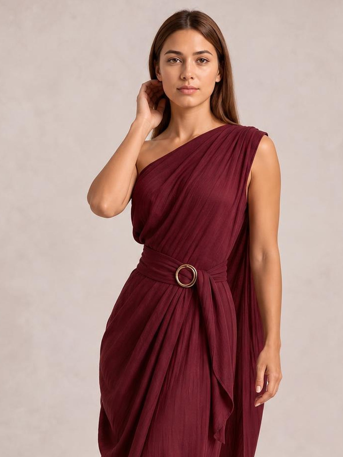 Model poses with one hand near her hair, wearing a burgundy one-shoulder draped maxi dress with a gathered waist and round buckle.