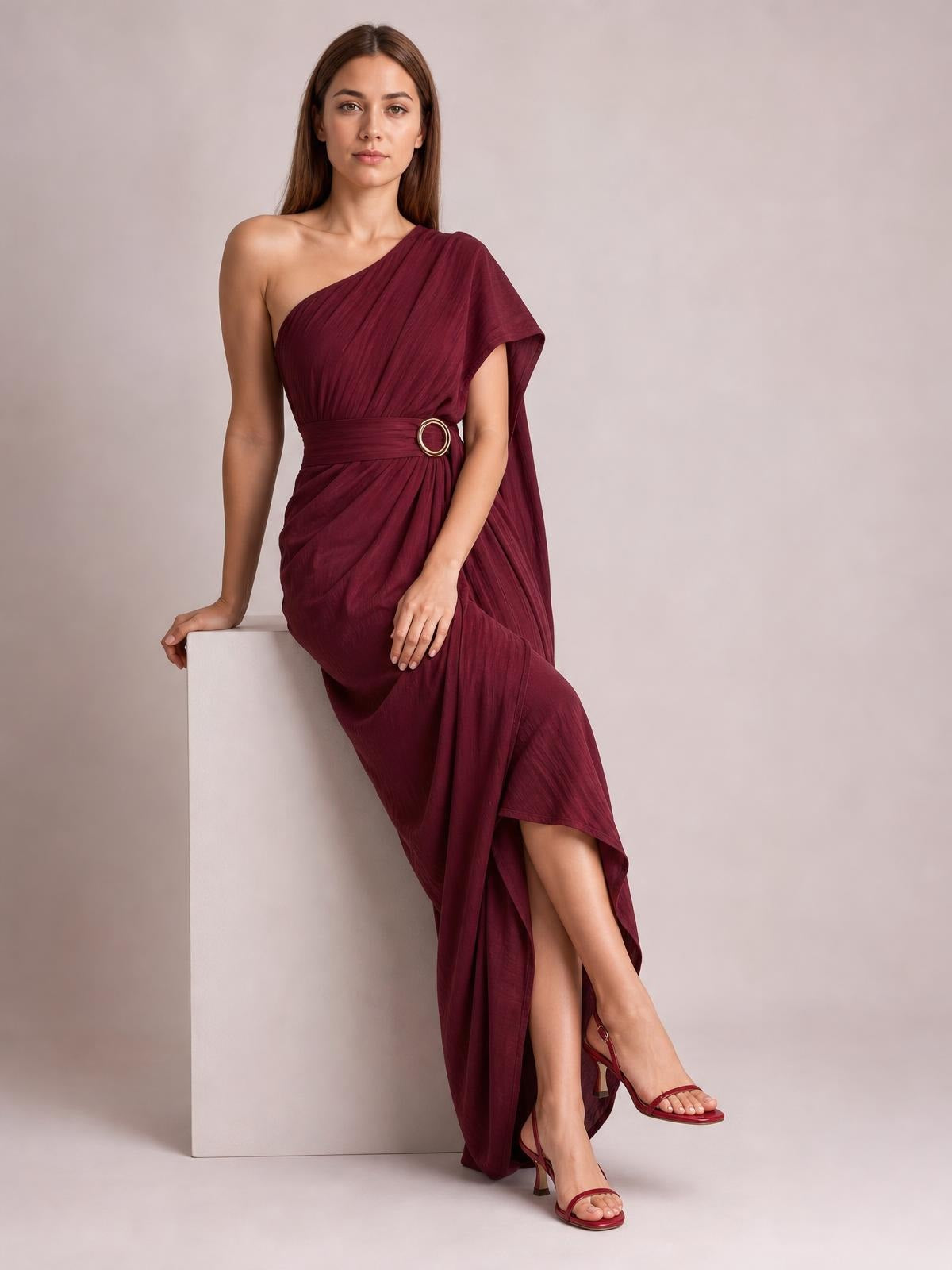 Model leans against a pedestal in a burgundy one-shoulder draped maxi dress with gathered fabric, a cinched waist buckle, and a flowing asymmetrical skirt.