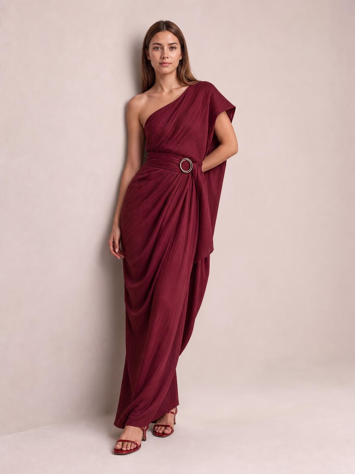 Model poses in a burgundy one-shoulder draped maxi dress with a flowing floor-length silhouette and circular waist buckle.