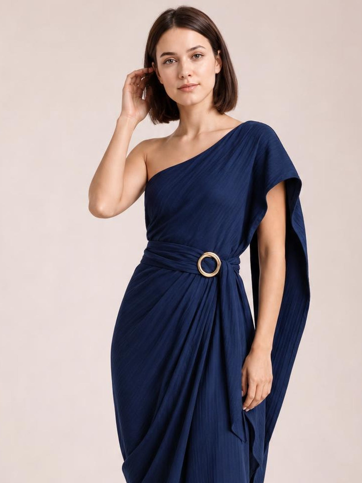 A model poses in a navy one-shoulder draped maxi dress with a gathered waist, circular buckle, flowing pleats, and an elegant asymmetric silhouette.
