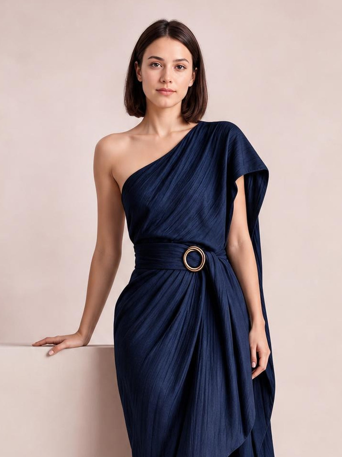 A model poses in a navy one-shoulder draped maxi dress with a gathered waist, circular buckle, flowing silhouette, and asymmetric sleeve.