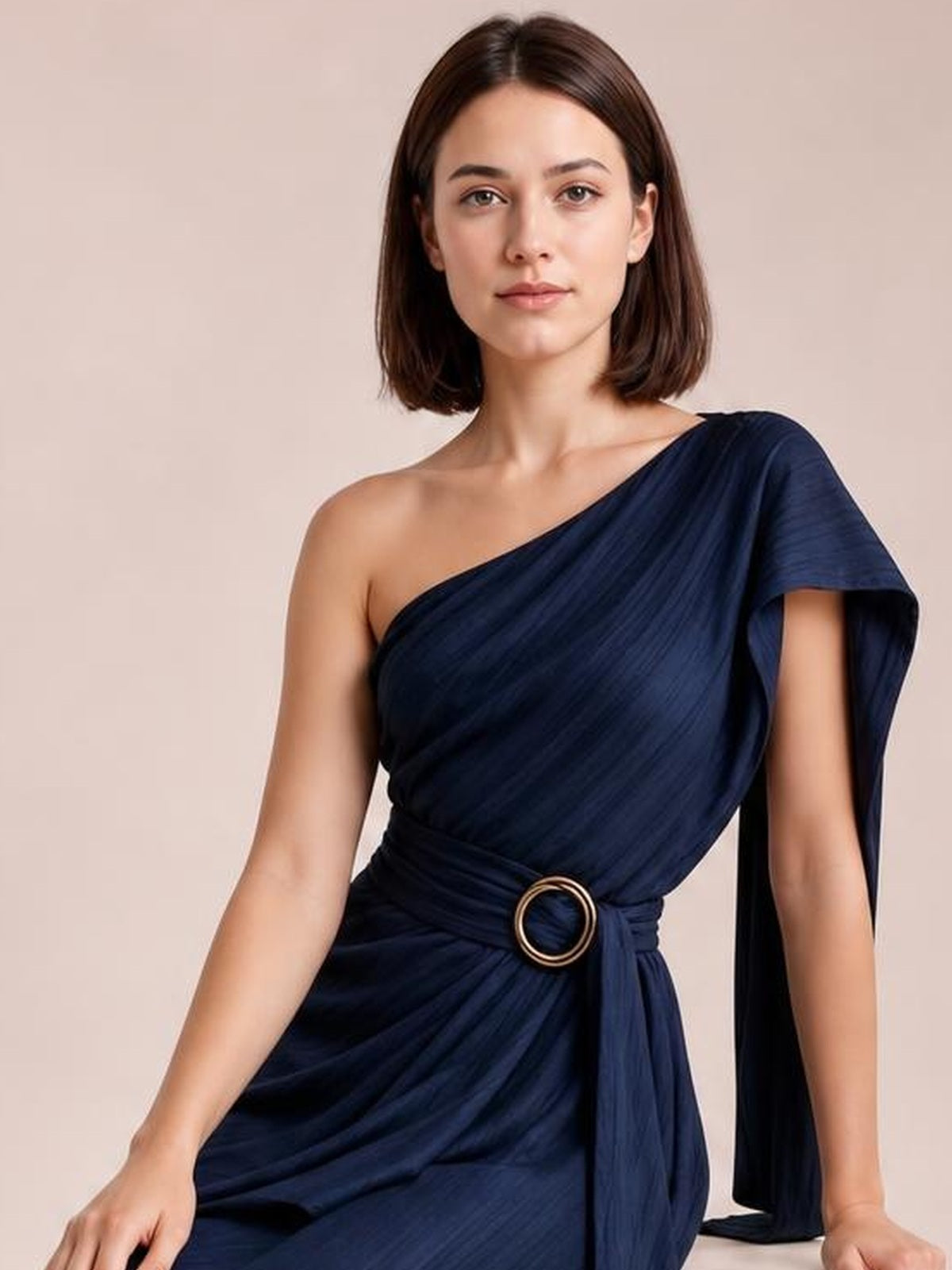 A model poses in a navy one-shoulder draped maxi dress with gathered fabric, a defined waist, and a round buckle detail.