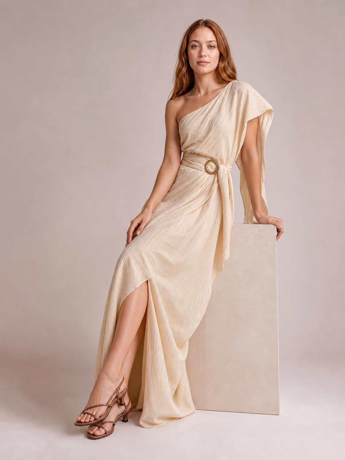Model poses seated in a cream one-shoulder draped maxi dress with a flowing silhouette, gathered waist, circular buckle, and cascading skirt.