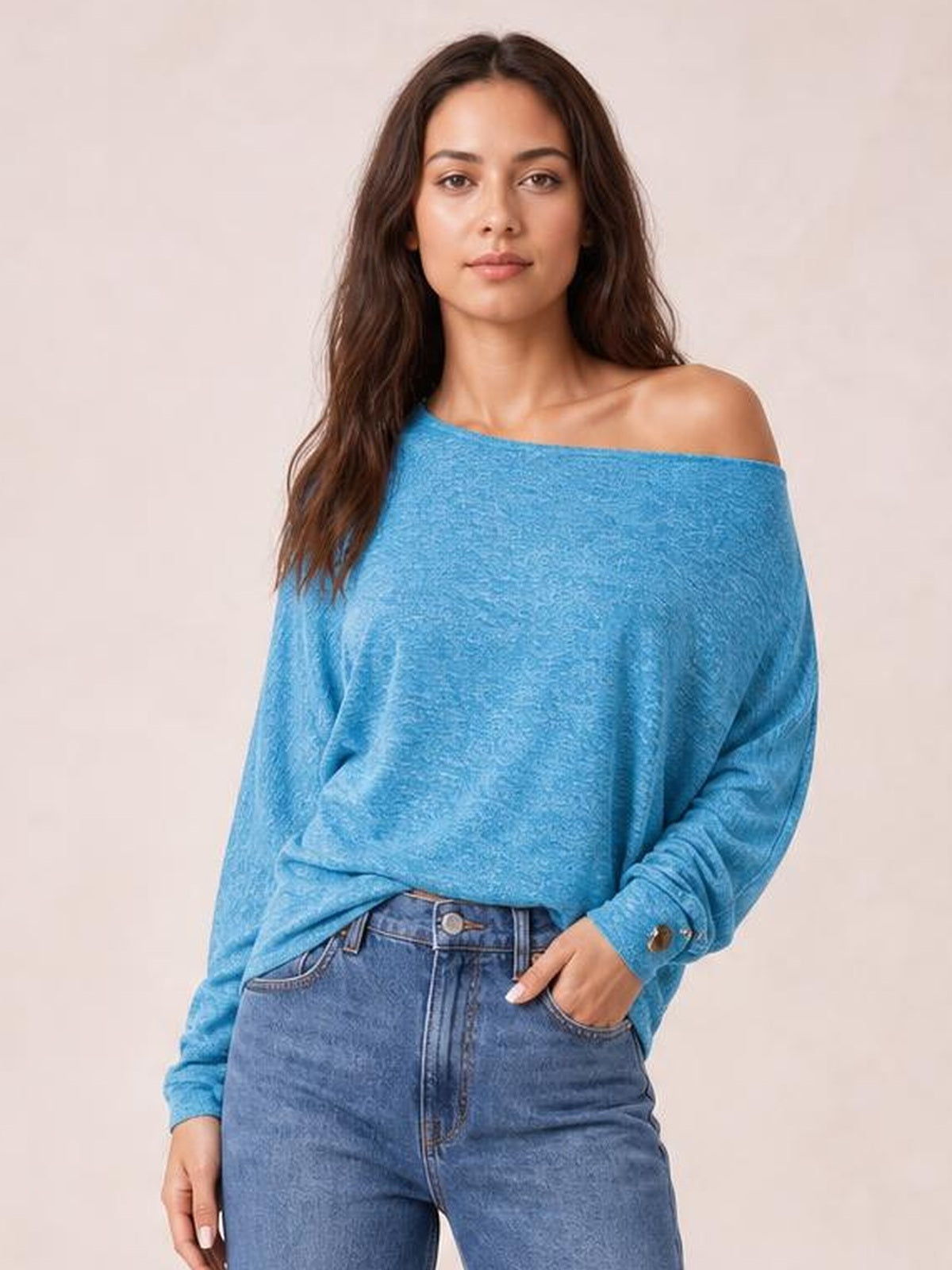 Model wearing a relaxed blue one-shoulder long-sleeve T-shirt with an exposed shoulder, draped silhouette, and decorative buttoned cuff.