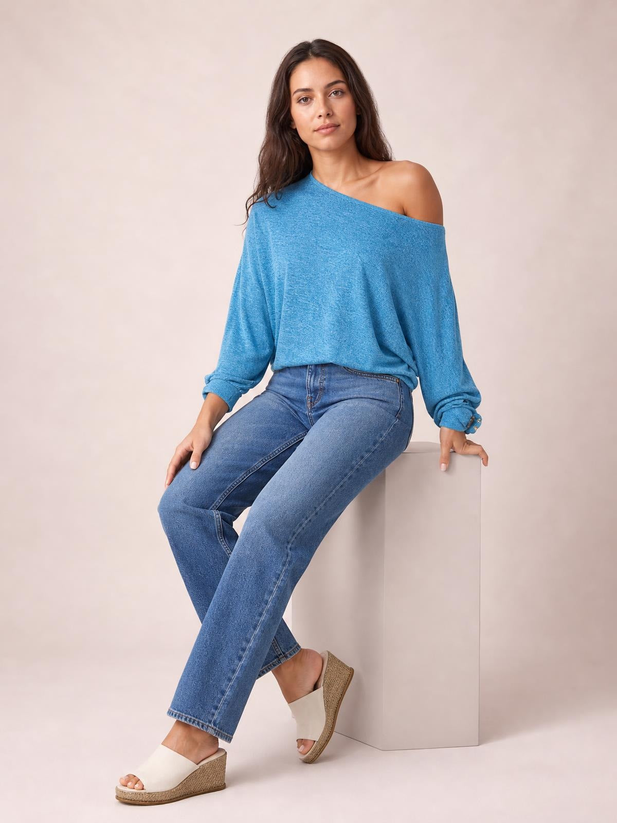 A seated model wears a blue one-shoulder long-sleeve T-shirt with a relaxed silhouette, gathered cuffs, and a decorative button, styled with crossed legs.