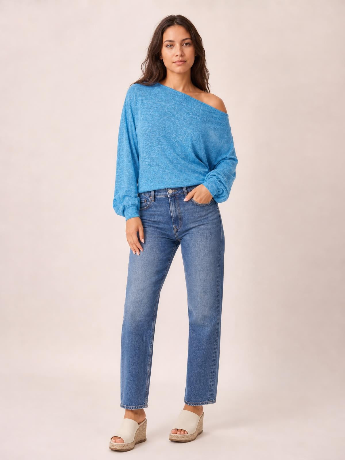 Model poses in a blue one-shoulder long-sleeve T-shirt with a draped neckline and gathered cuffs, paired with straight-leg jeans.