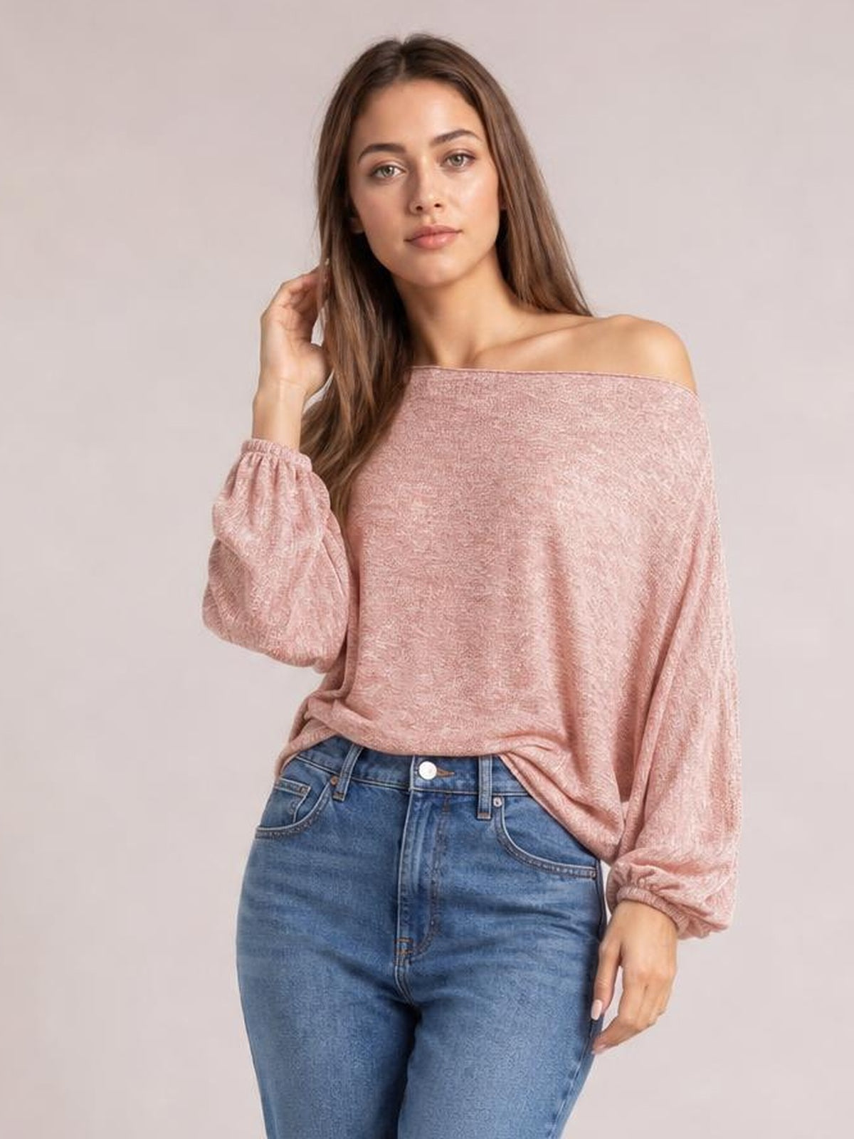 Model wearing a blush pink one-shoulder long-sleeve T-shirt with a wide neckline, relaxed silhouette, and gathered cuffs while touching her hair.