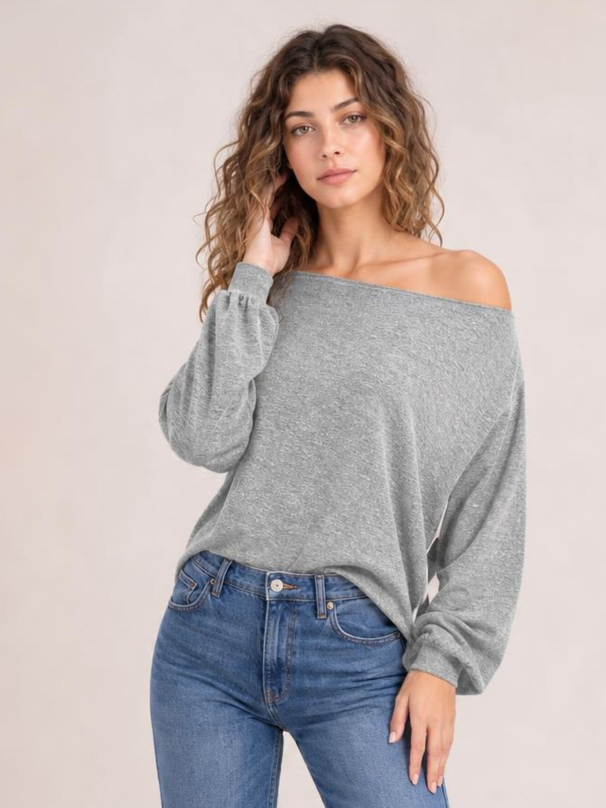 A model poses in a gray one-shoulder long-sleeve T-shirt with a wide neckline and relaxed silhouette, one hand in her hair and the other at her side.