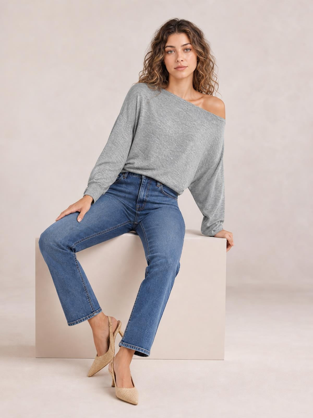 A woman seated in a relaxed pose wears a gray one shoulder long sleeve T-shirt with a draped silhouette and exposed shoulder.