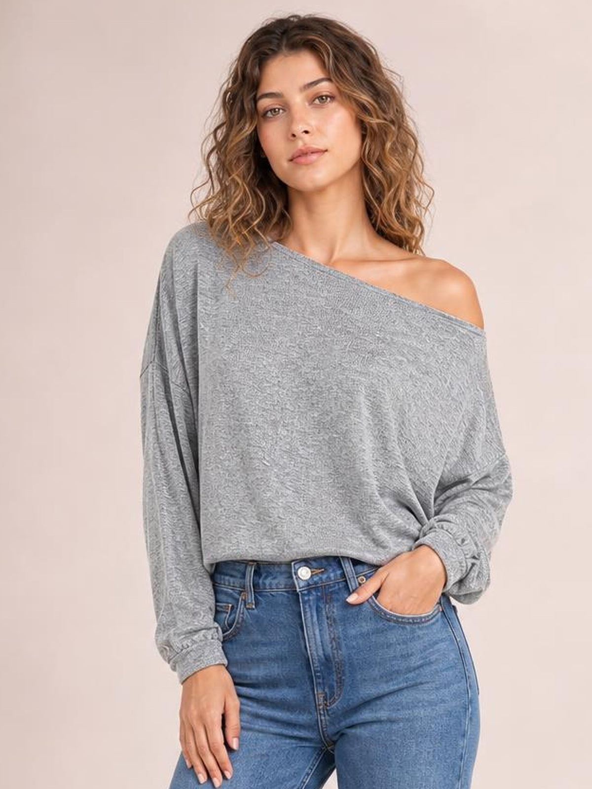 Model wears a gray one-shoulder long-sleeve T-shirt with a relaxed silhouette, wide neckline, gathered cuffs, and a hand resting in her jeans pocket.