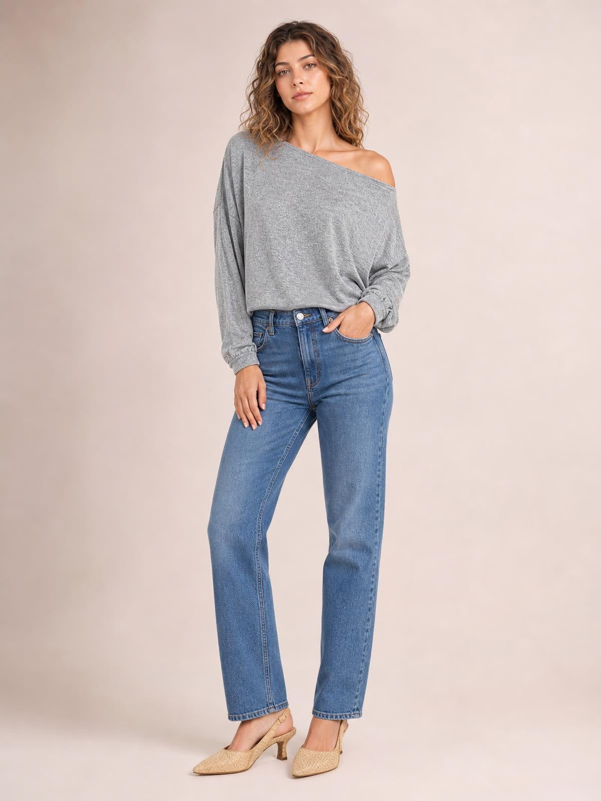 Model wears a gray one shoulder long sleeve T-shirt with a relaxed silhouette, angled neckline, and gathered sleeve detail while standing with one hand in her pocket.