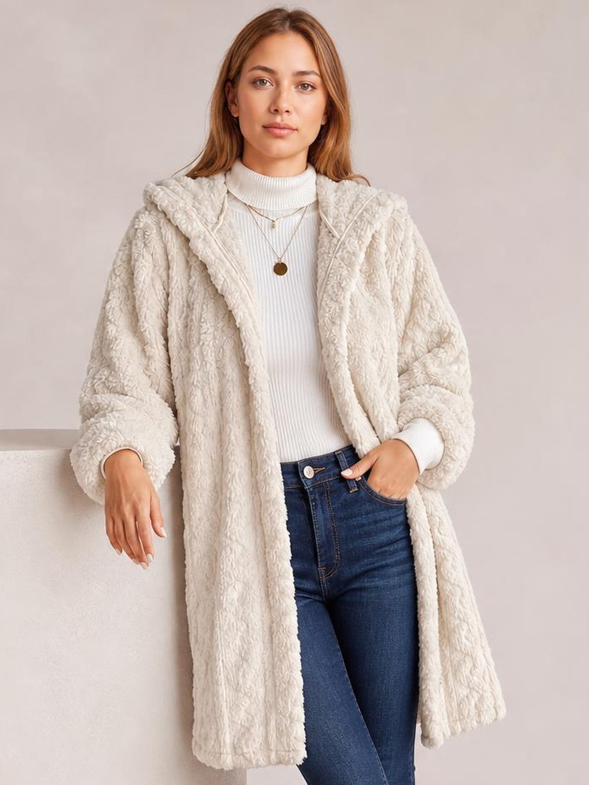 Model wears a long cream fuzzy hooded jacket with open front, long sleeves, plush textured construction, and a relaxed silhouette over a white sweater and jeans.