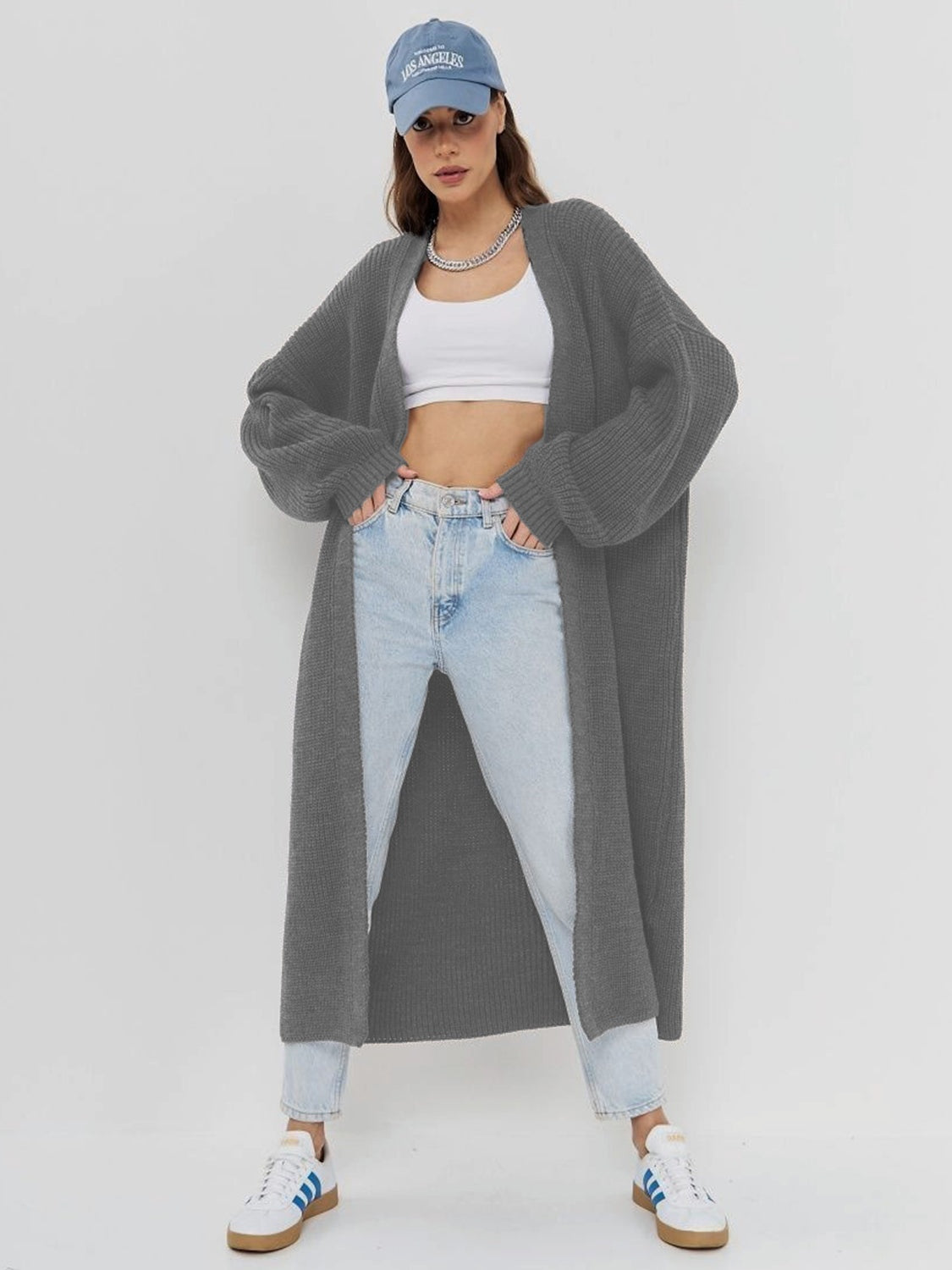 Model poses with hands in the pockets of a gray open-front longline cardigan featuring long sleeves, ribbed texture, and a relaxed ankle-length silhouette.