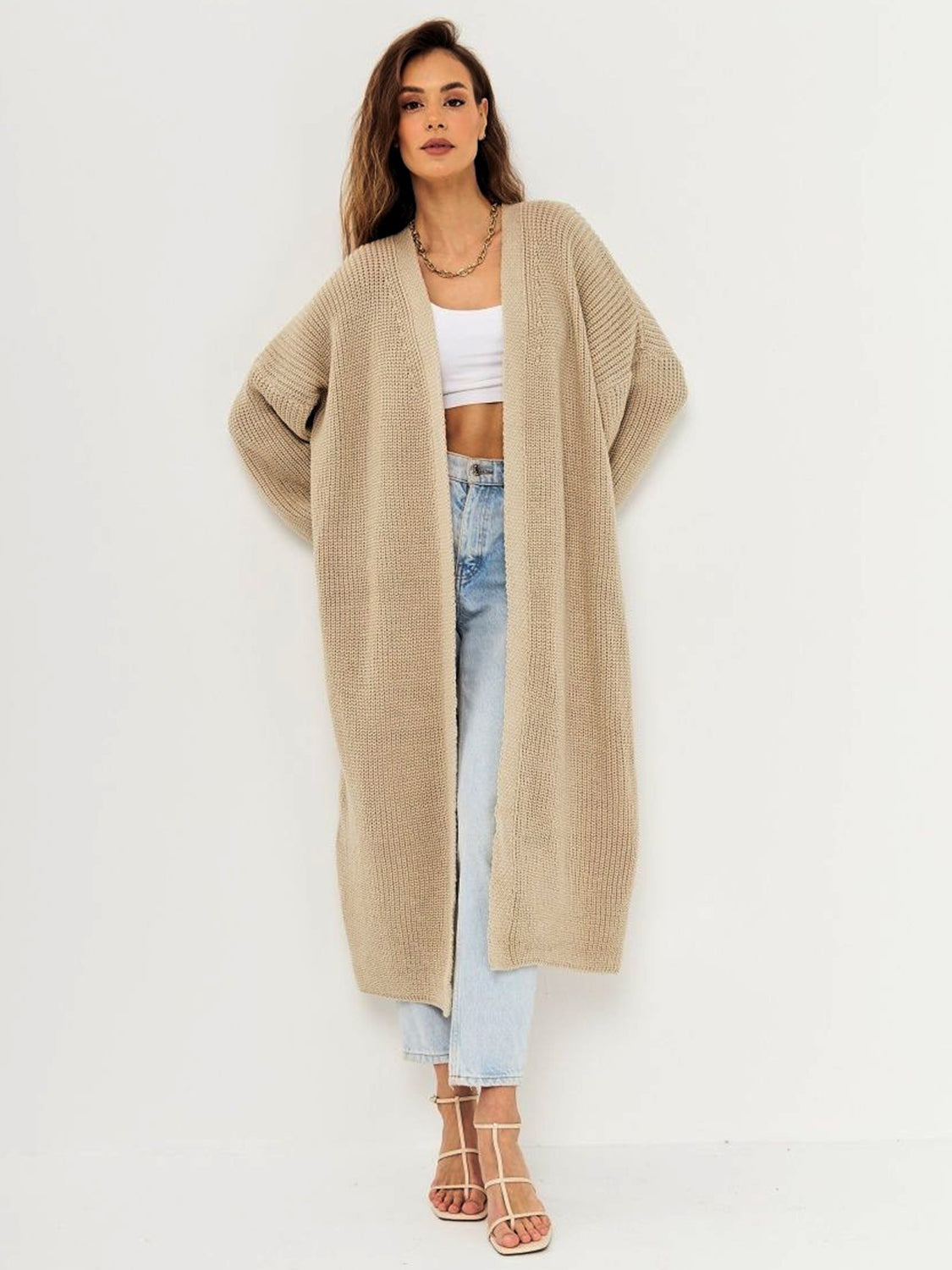 Model poses in a beige open-front long-sleeve longline cardigan with a relaxed silhouette, textured knit construction, and extended hem over jeans.