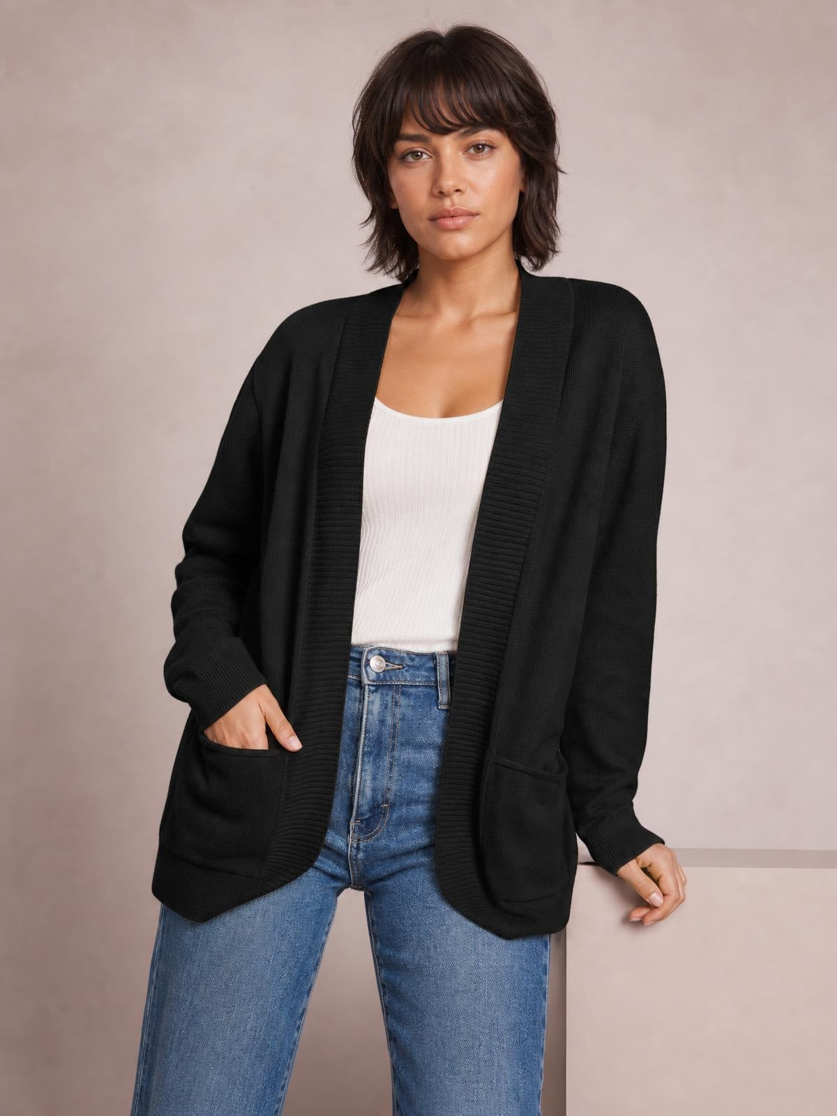 A woman wears a black open front long sleeve sweater cardigan with ribbed trim, a relaxed silhouette, and patch pockets over a white top and jeans.