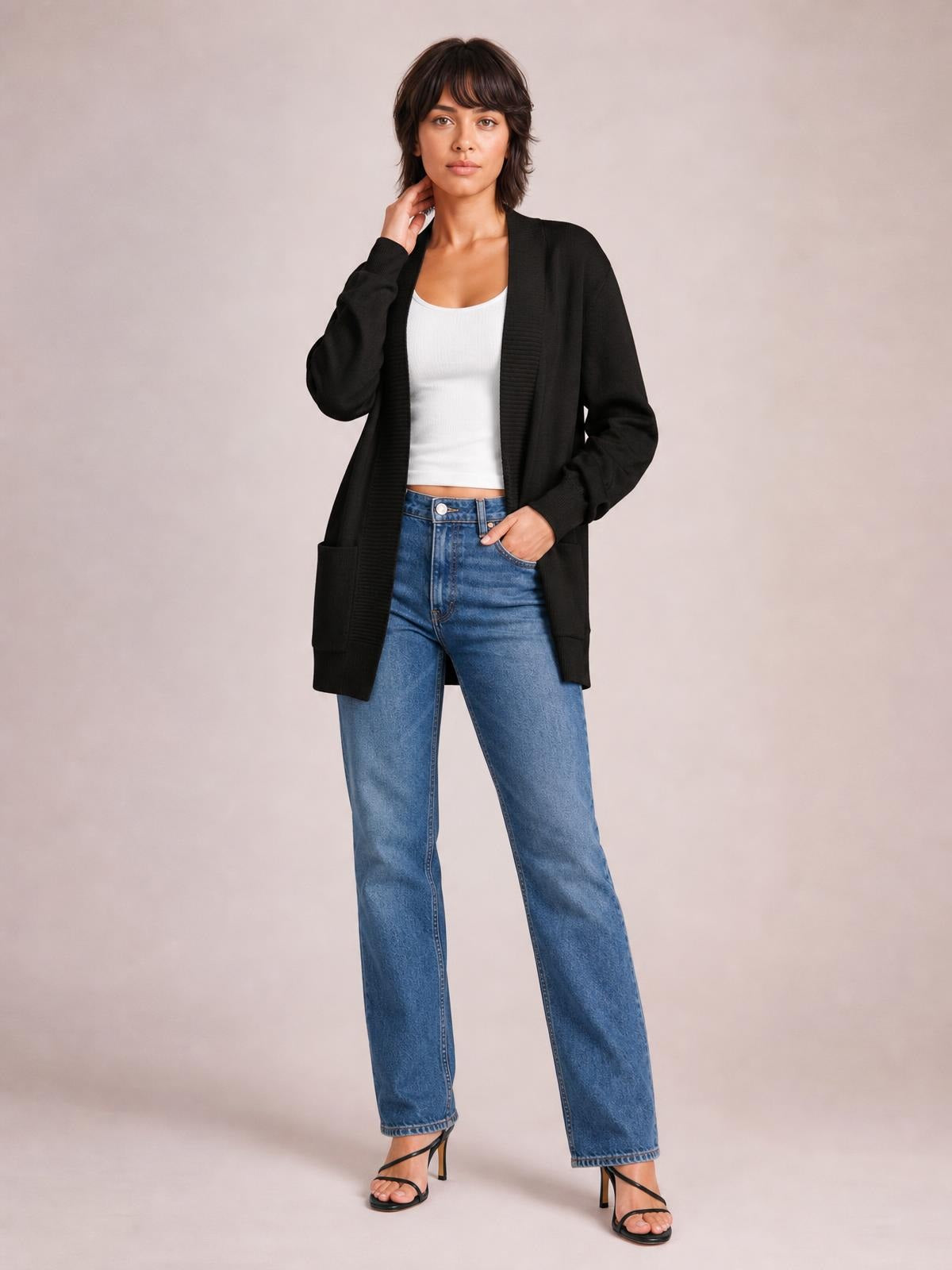 Model poses in a black open-front long sleeve sweater cardigan with ribbed trim, a long relaxed silhouette, and front pockets over a white top.