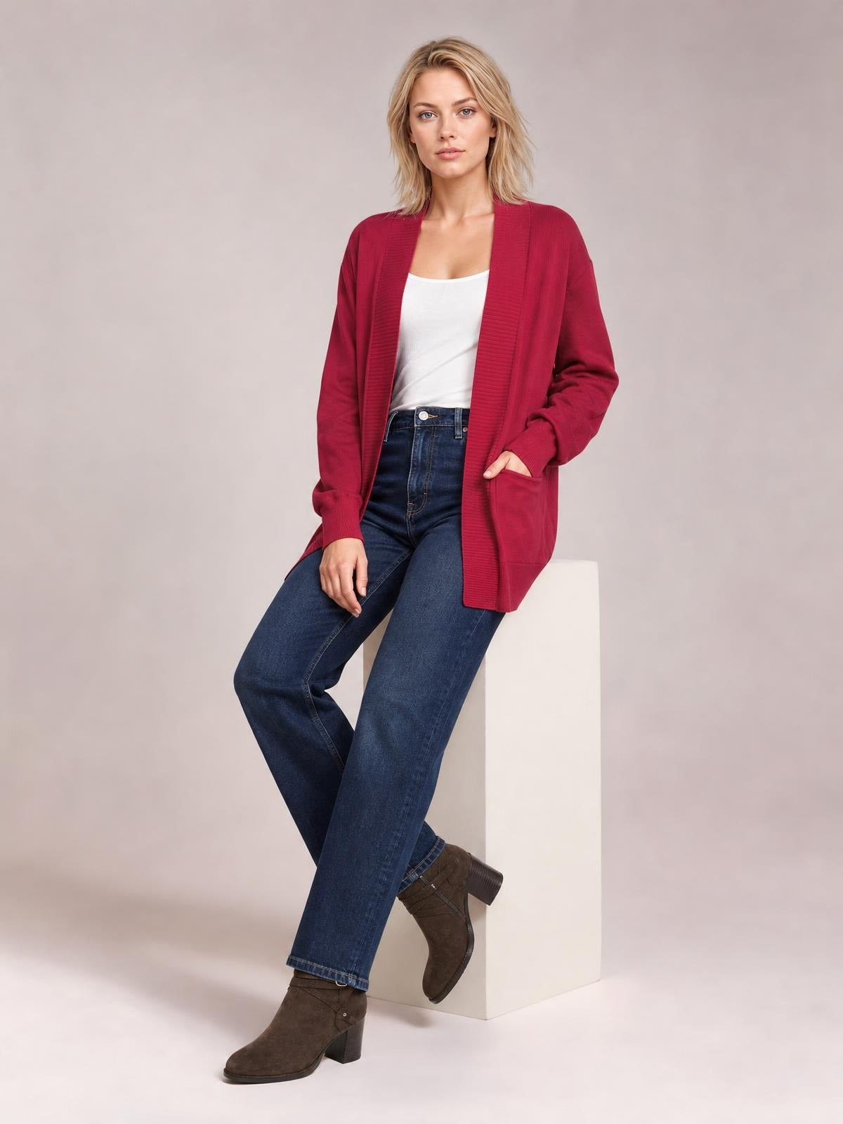 Model seated in a relaxed pose wearing a burgundy open front long sleeve sweater cardigan with ribbed trim, patch pockets, and a long straight silhouette.
