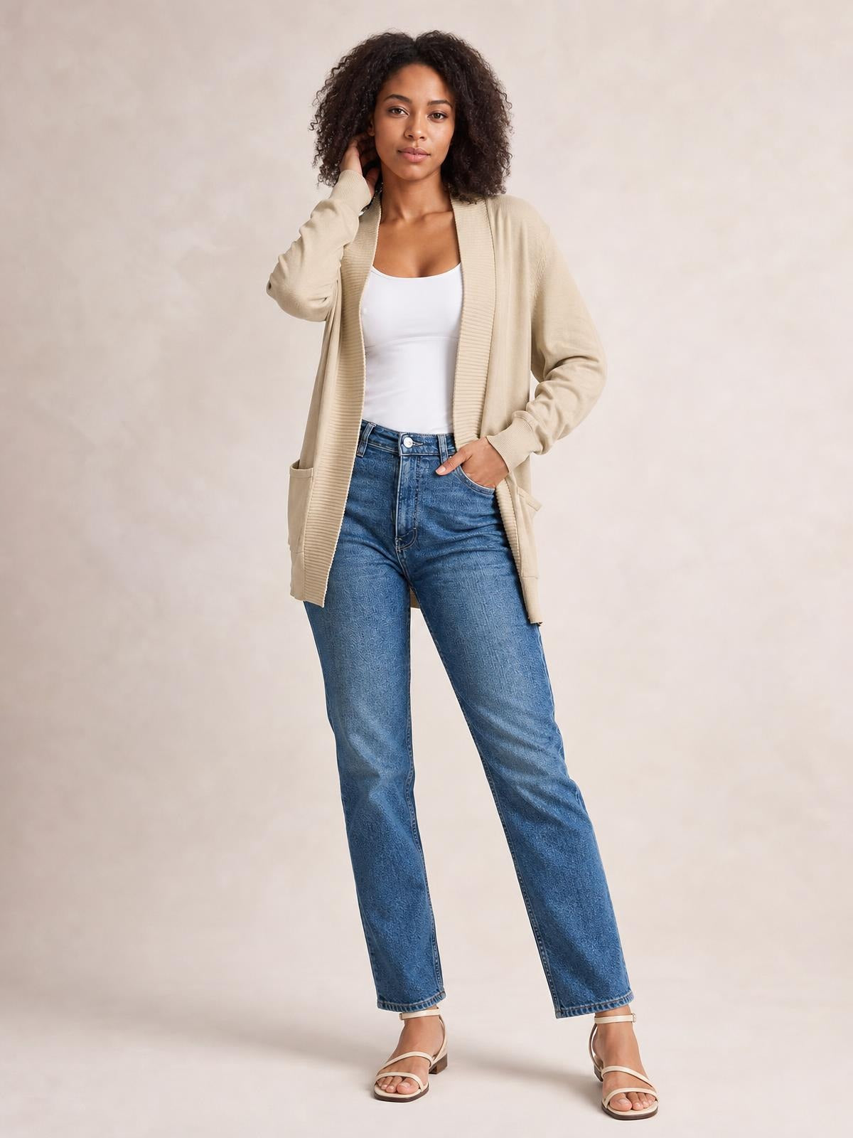 A model wears a beige open-front long-sleeve sweater cardigan with a relaxed long silhouette and patch pockets over a white top and blue jeans.