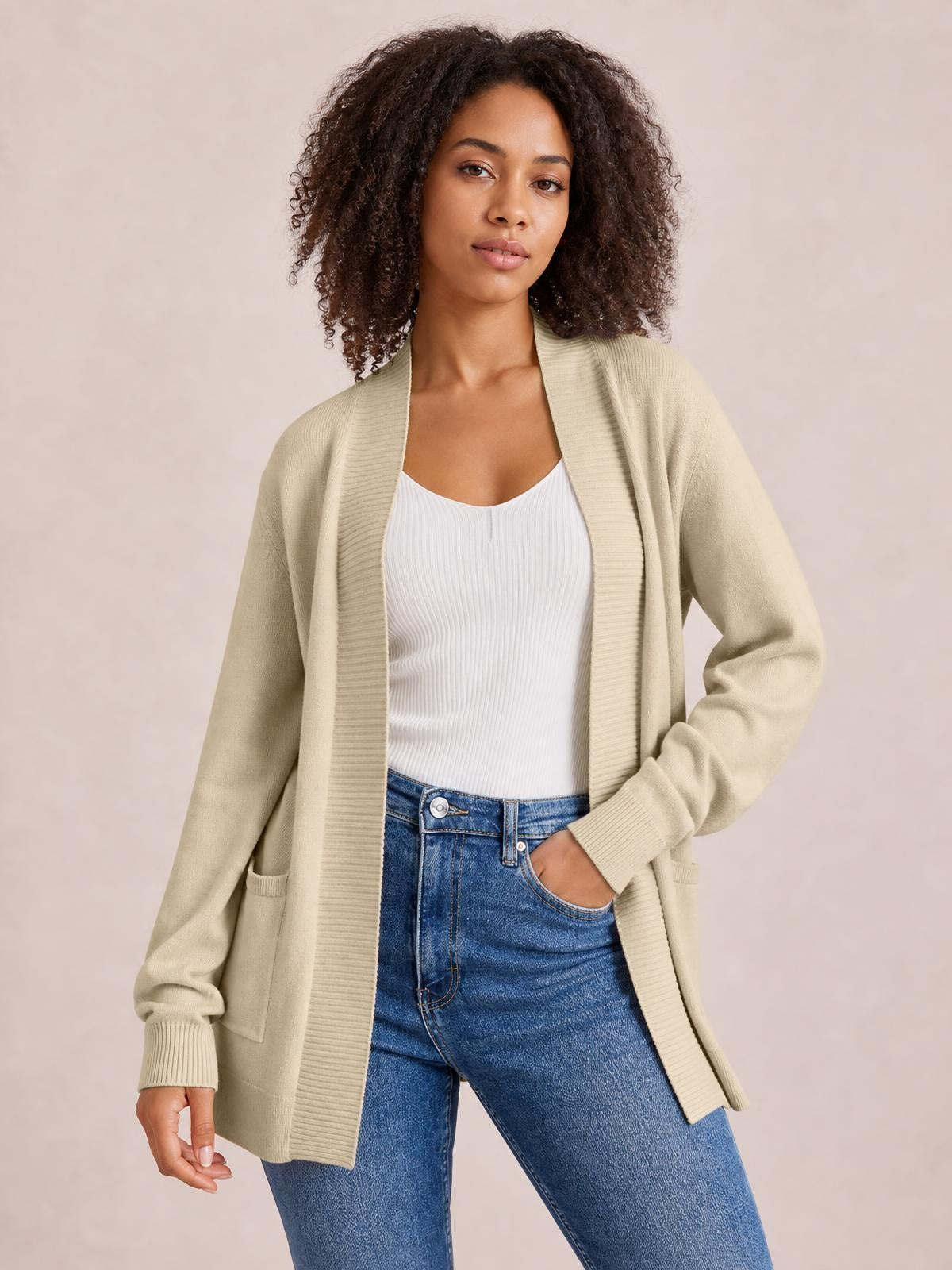 A model wears a beige open-front long-sleeve sweater cardigan with ribbed trim, front pockets, and a relaxed long silhouette, posing with one hand in her jeans pocket.