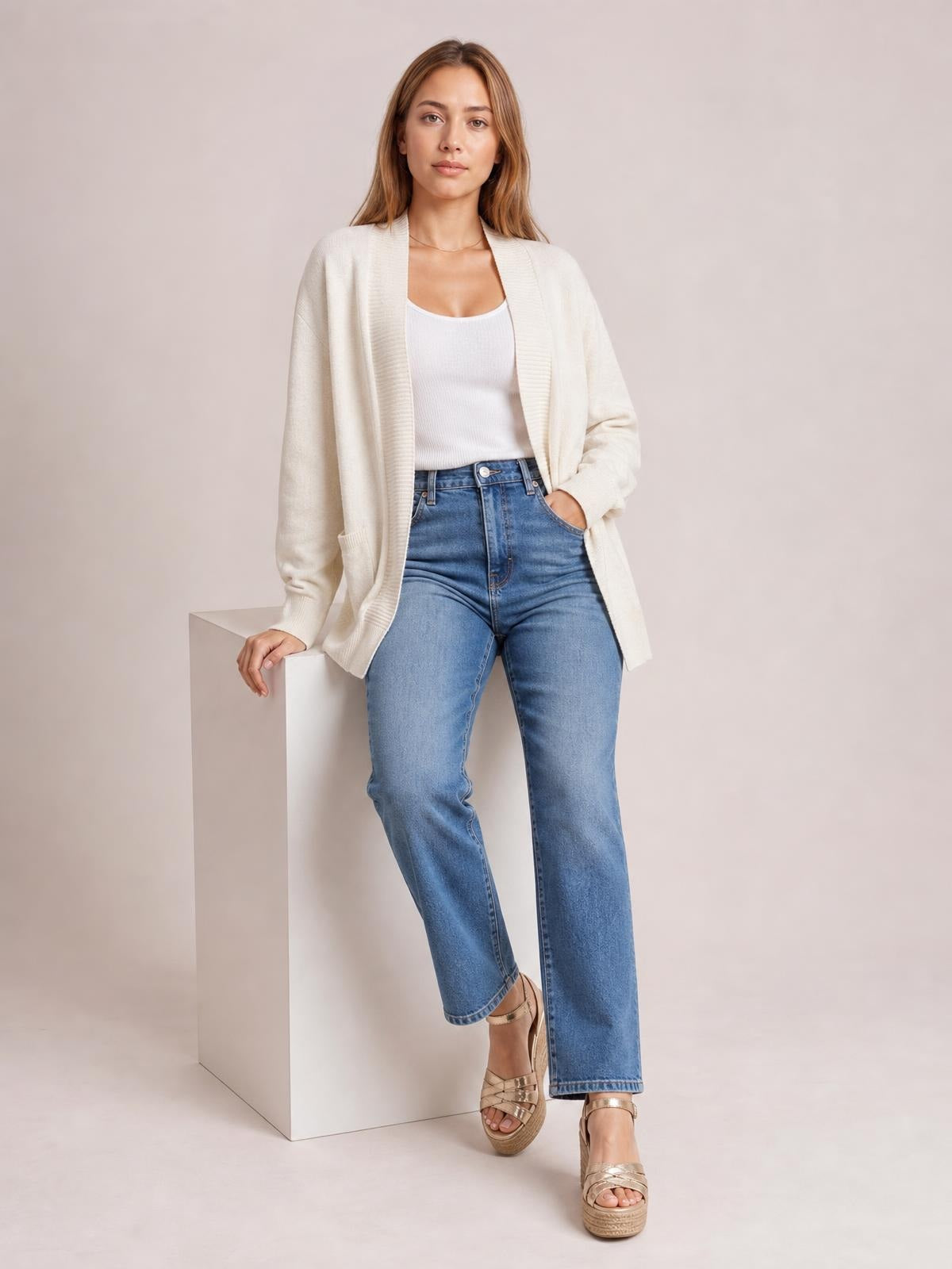 A model poses in a cream open-front long sleeve sweater cardigan with a relaxed silhouette, ribbed trim, and visible patch pockets over a white top.