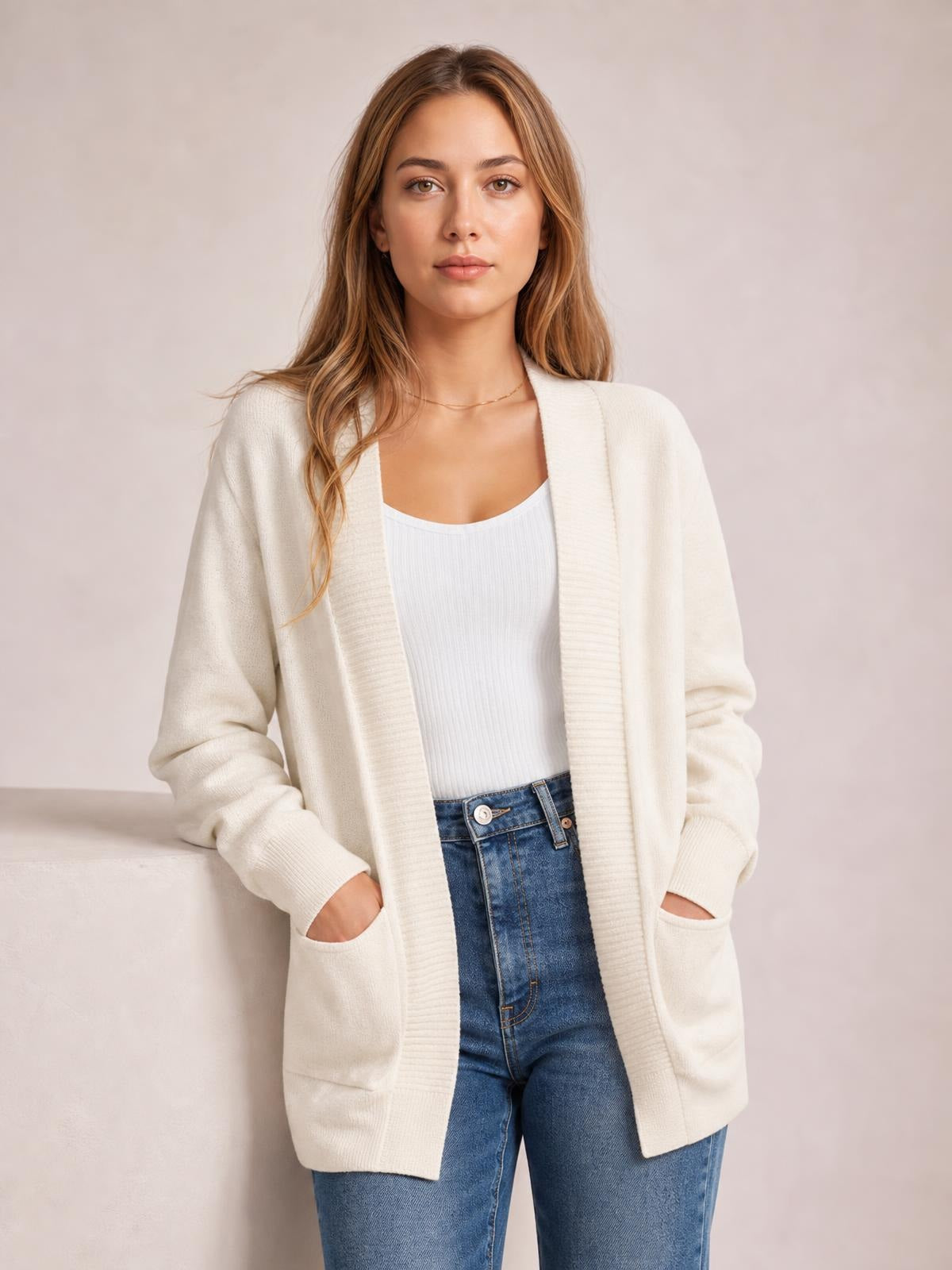Model wears a cream open-front long-sleeve sweater cardigan with a relaxed long silhouette, ribbed trim, and two front pockets over a white top.