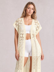 Model standing in a cream open-front short-sleeve openwork cover-up with a flowy long silhouette and delicate ruffled trim over white activewear.