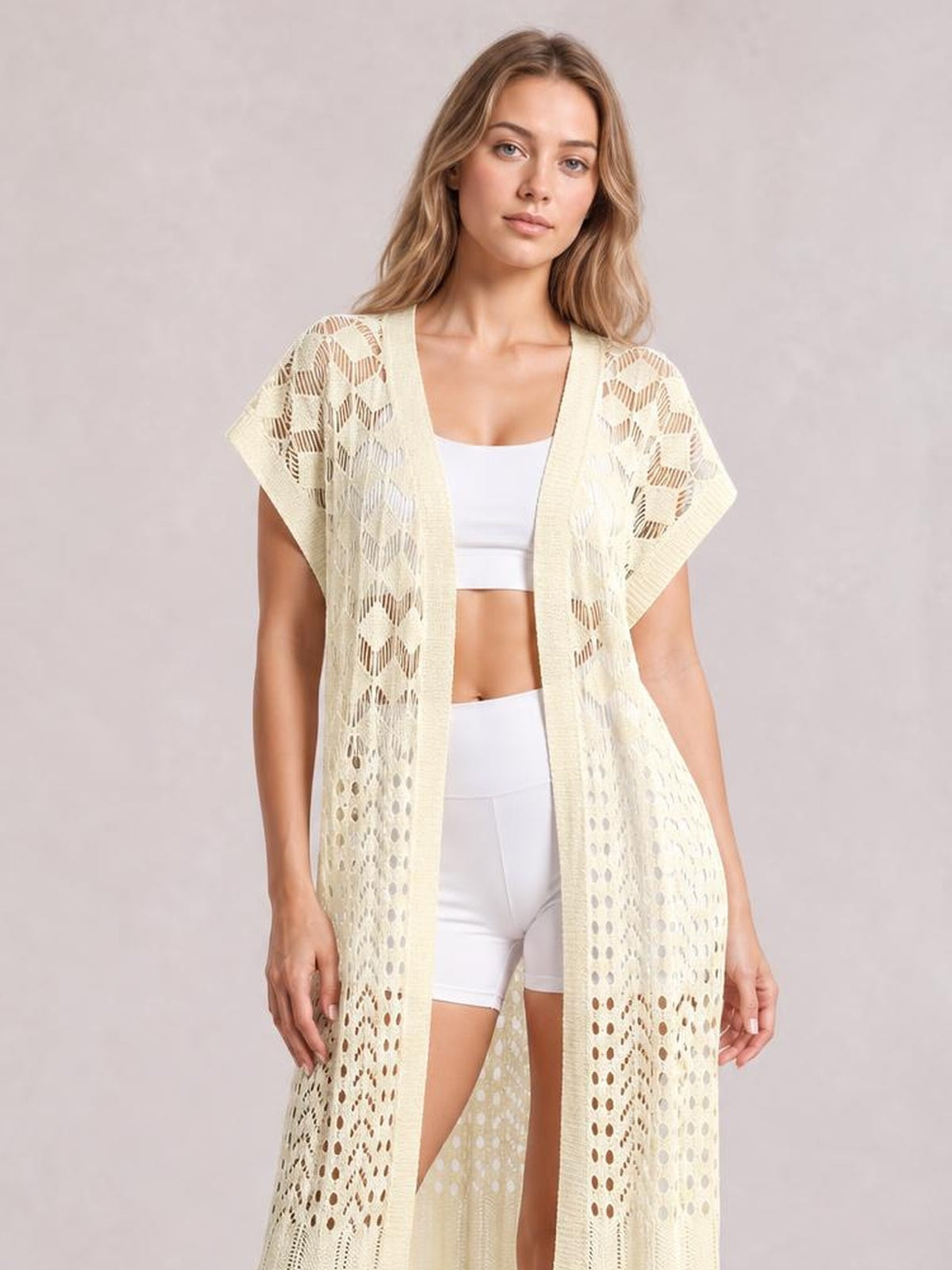 Model standing in a cream open-front short-sleeve openwork cover-up with a flowy long silhouette and delicate ruffled trim over white activewear.