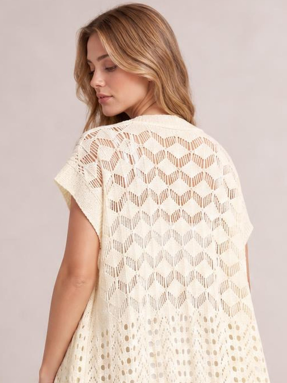 Model turns to show a cream openwork cover-up with short sleeves, a flowy silhouette, geometric cutouts, and delicate ruffled detailing.