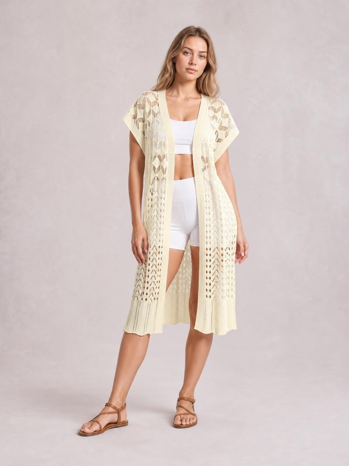 Model wears a cream open-front short-sleeve openwork cover-up with delicate ruffled details and a long, flowy silhouette over white separates.