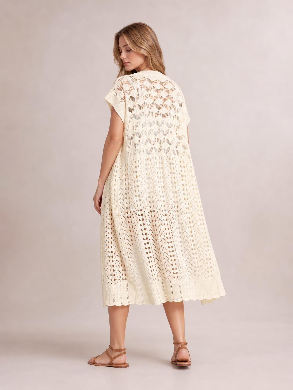 A model turns to show an ivory open front short sleeve openwork cover-up with a flowy silhouette, patterned crochet-style construction, and scalloped hem.