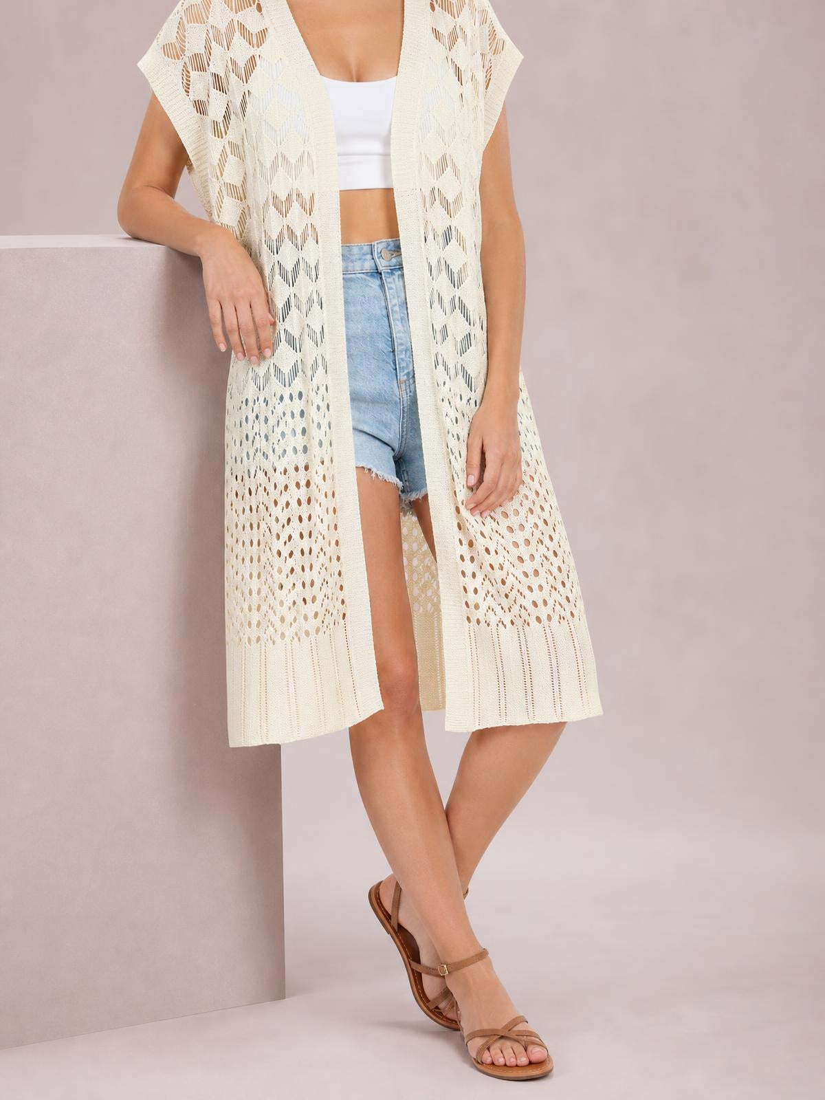 A model wears a cream open-front short-sleeve openwork cover-up with a flowy long silhouette and patterned cutout construction, posing with crossed legs.